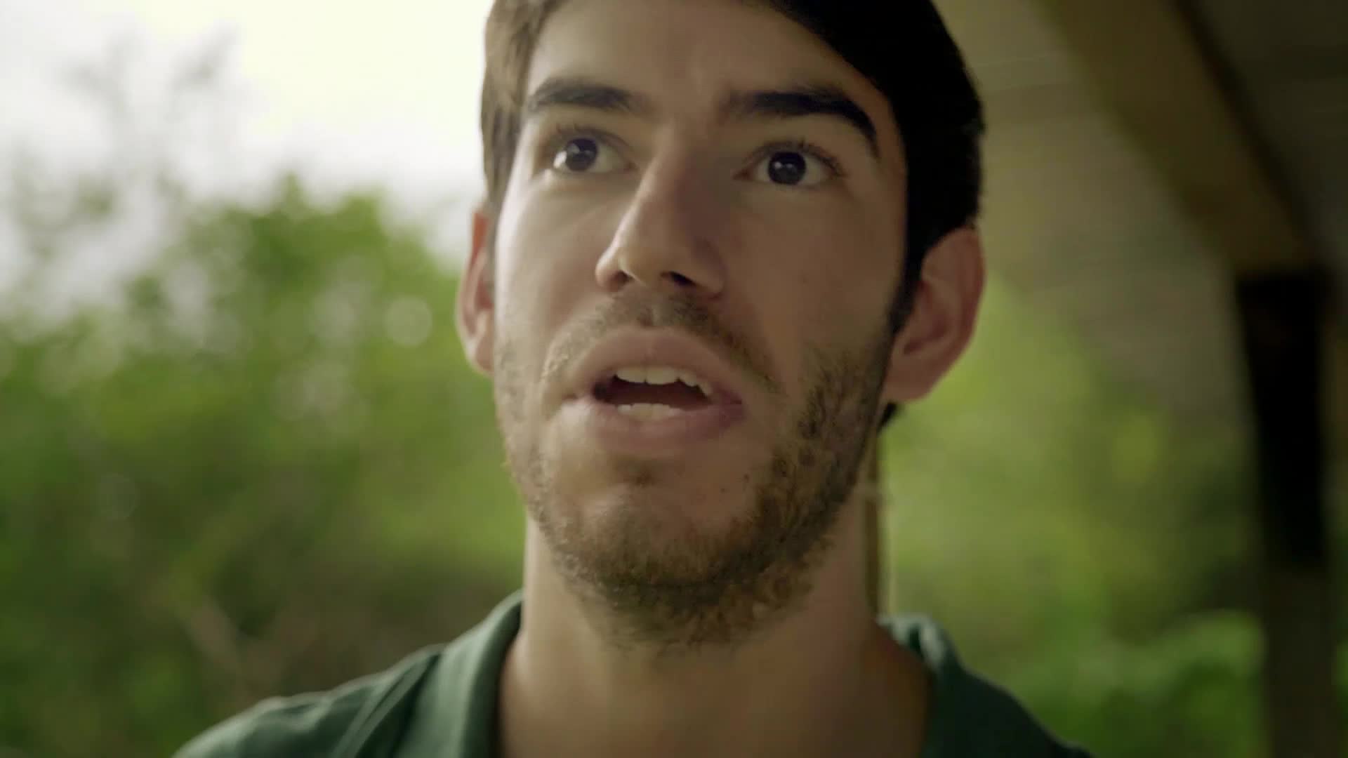 A young man with a beard speaks urgently, his eyes wide. The background is a soft blur of green trees and a wooden overhang.