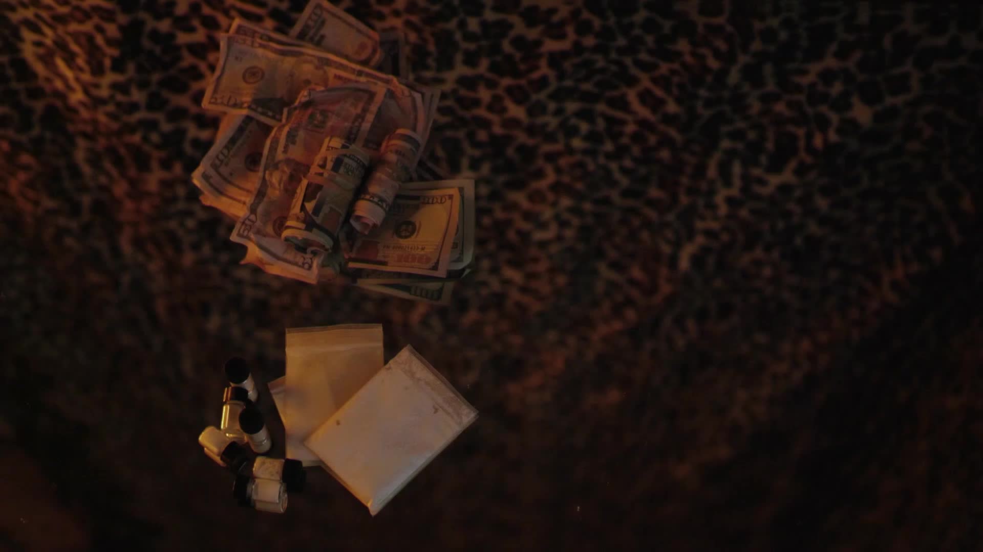 A pile of crumpled US dollars sits on a leopard-print surface. Beside the money, small vials and plastic baggies of white powder are arranged.