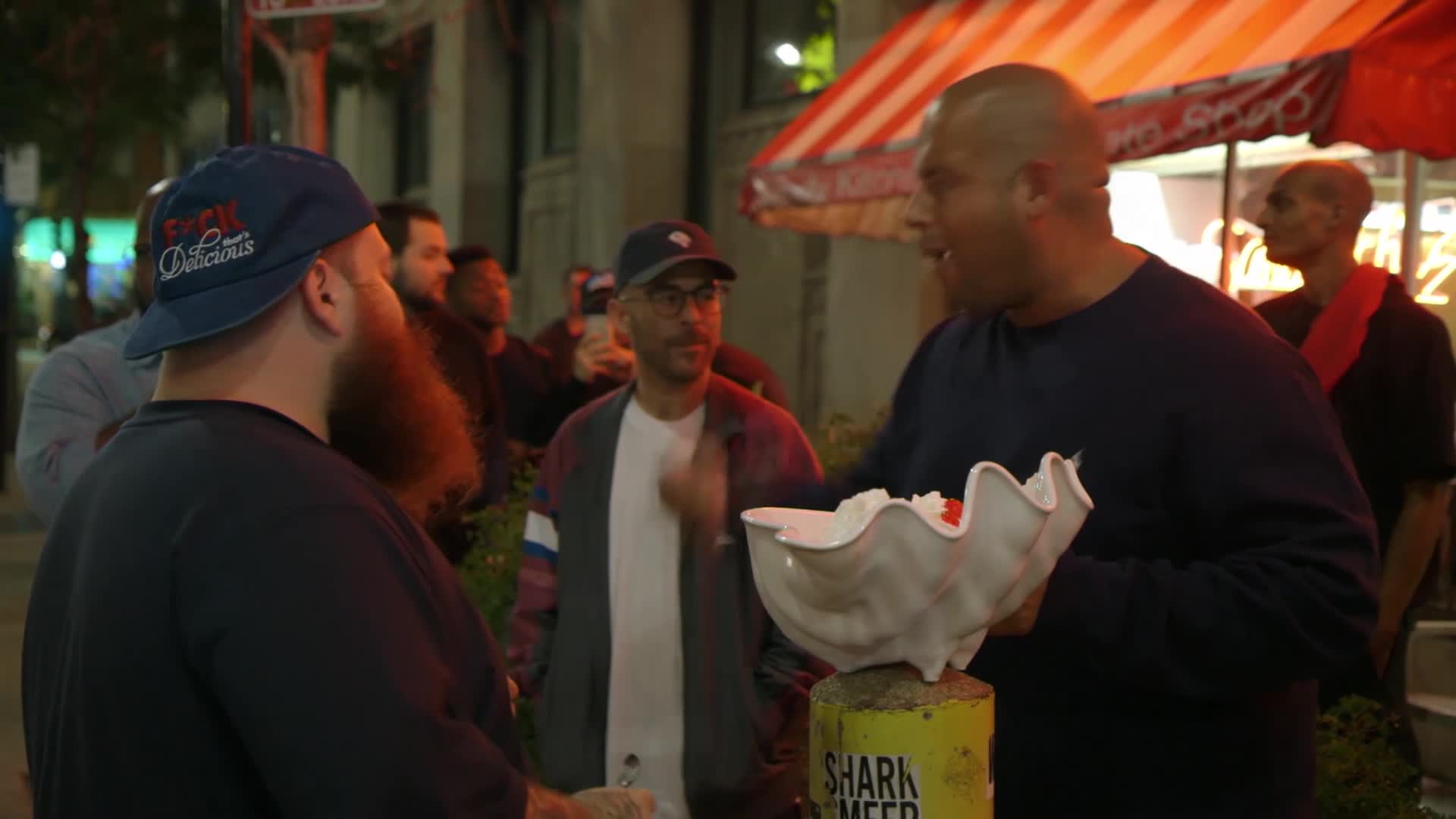 A man with a full beard and a blue cap holds a large white bowl filled with what looks like whipped cream and berries. He's talking animatedly to another man in a dark blue shirt, who is also looking at the bowl.