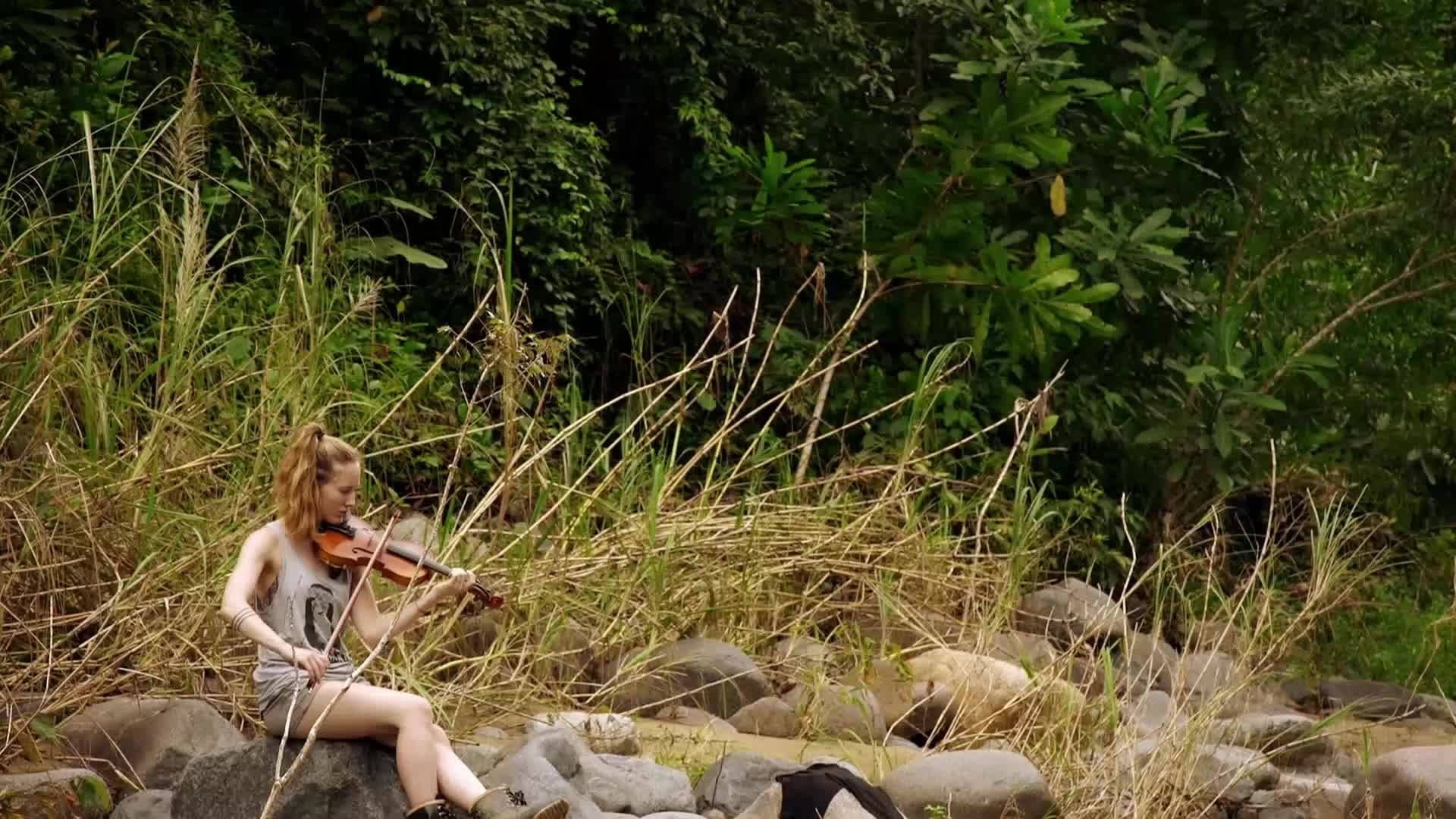 A young woman sits on a rock, playing a violin amidst tall grasses and dense foliage. The sound of her bow against the strings cuts through the quiet hum of the jungle.