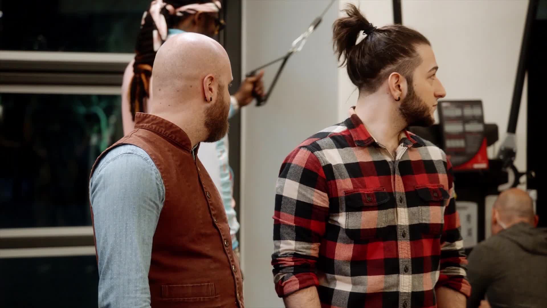 A man with a topknot and a red plaid shirt stands facing right, his gaze fixed on something out of frame. Behind him, a bald man in a denim shirt and brown vest turns his head, also looking off to the side.