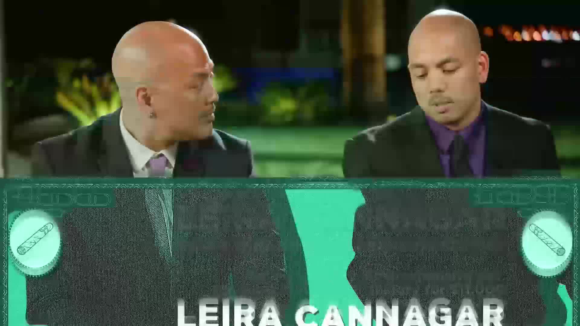 Two men in suits are seated, one looking off to the side while the other stares down. A graphic overlay with the name "LEIRA CANNAGAR" runs across the bottom of the screen.