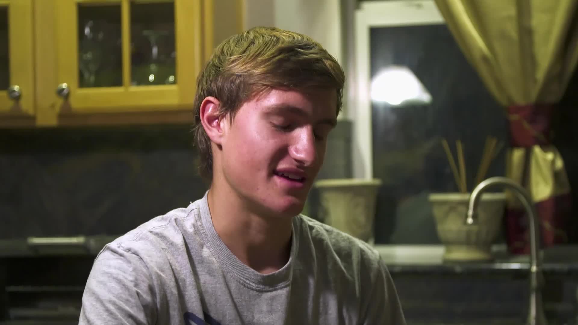 A young man with tousled brown hair speaks, his eyes cast down as if recalling a difficult moment. Behind him, a kitchen counter holds a faucet and some decorative pots, bathed in the dim light of a nearby window.