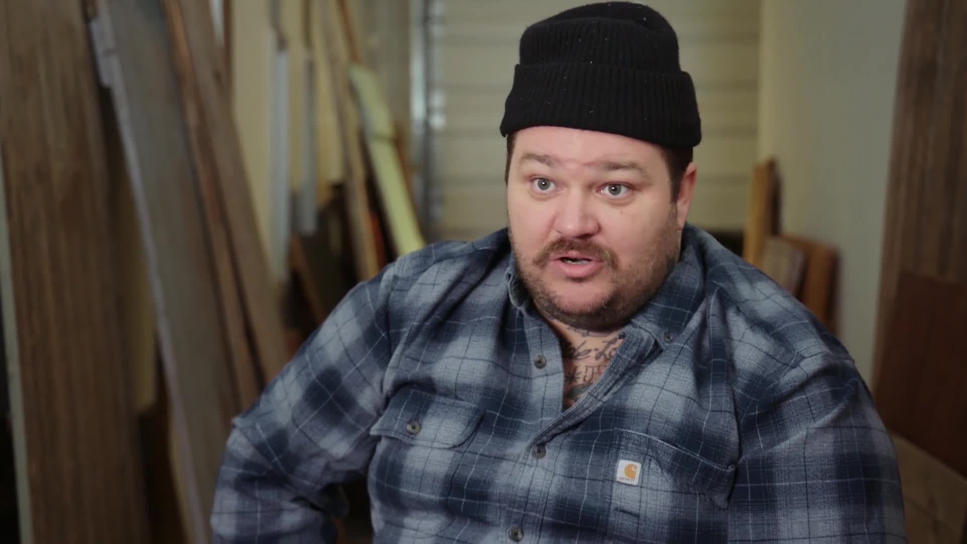 A man in a black beanie and a blue plaid shirt speaks directly to the camera. He's surrounded by stacks of lumber, suggesting a workshop setting.