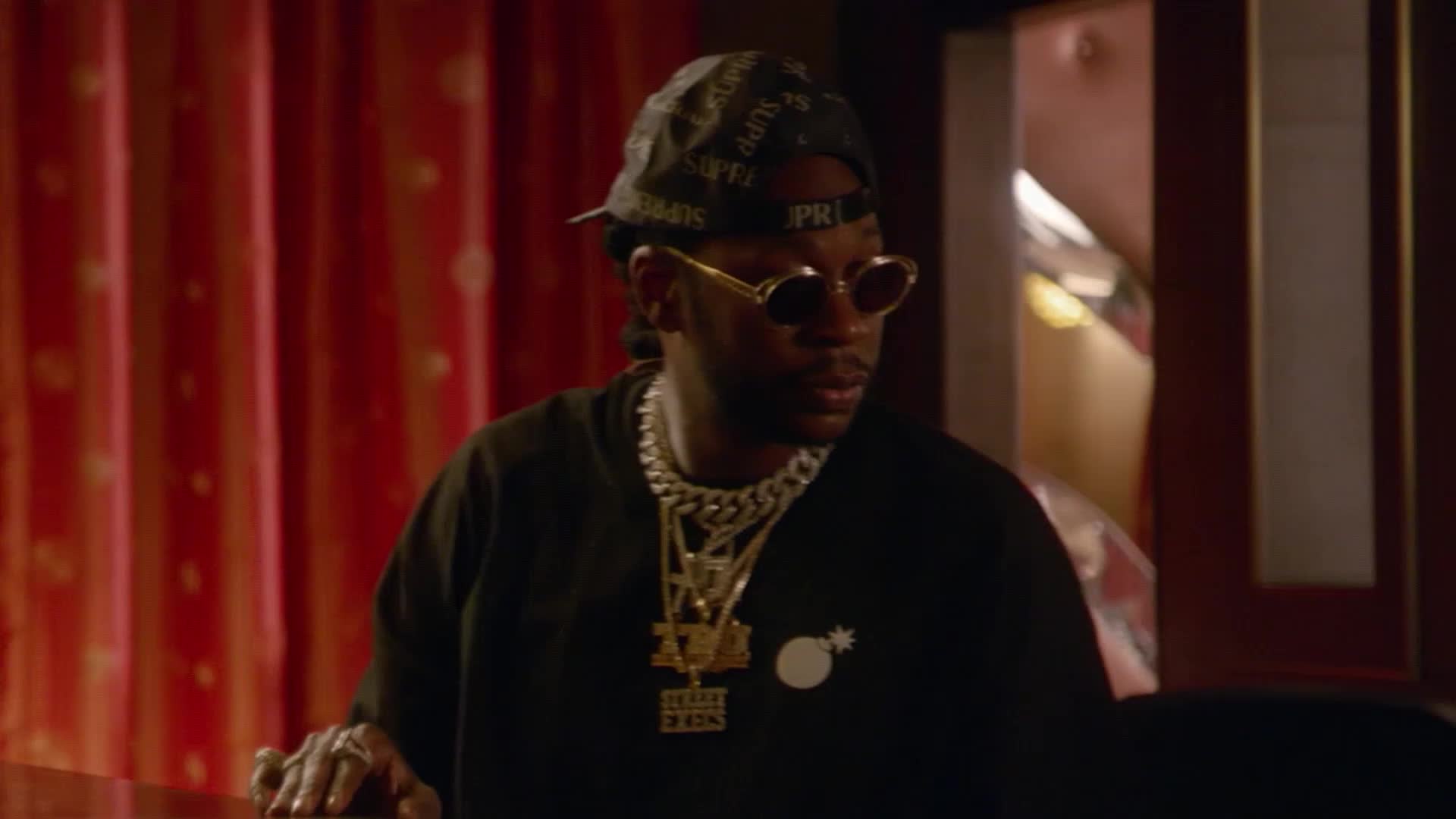 A man in a black t-shirt and a patterned bandana leans forward, his hand resting on a polished surface. He wears thick chains and oval sunglasses, his gaze fixed off-camera.