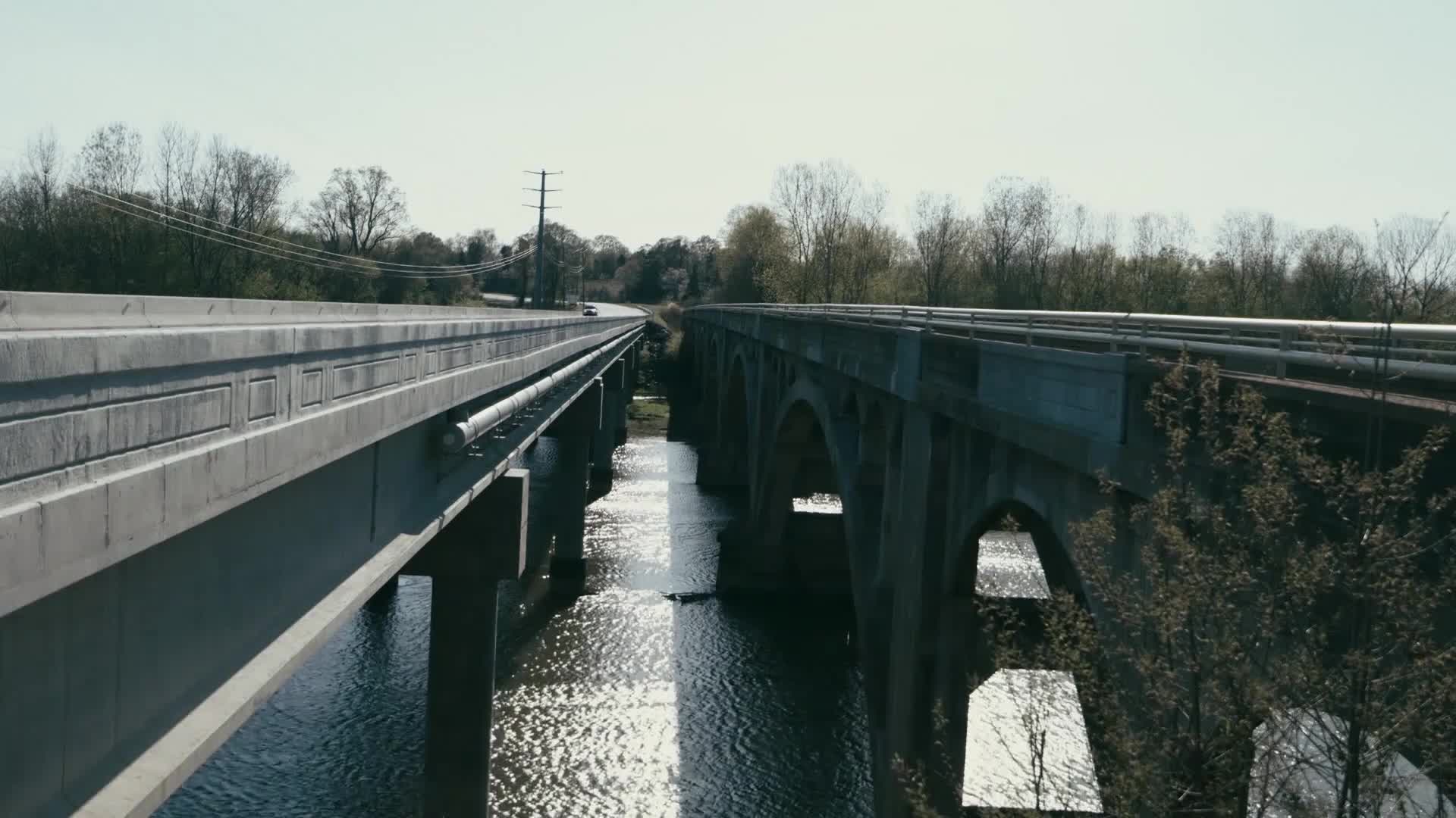 A lone car travels down the highway, crossing a wide river on a concrete bridge. The water below reflects the bright, clear sky.