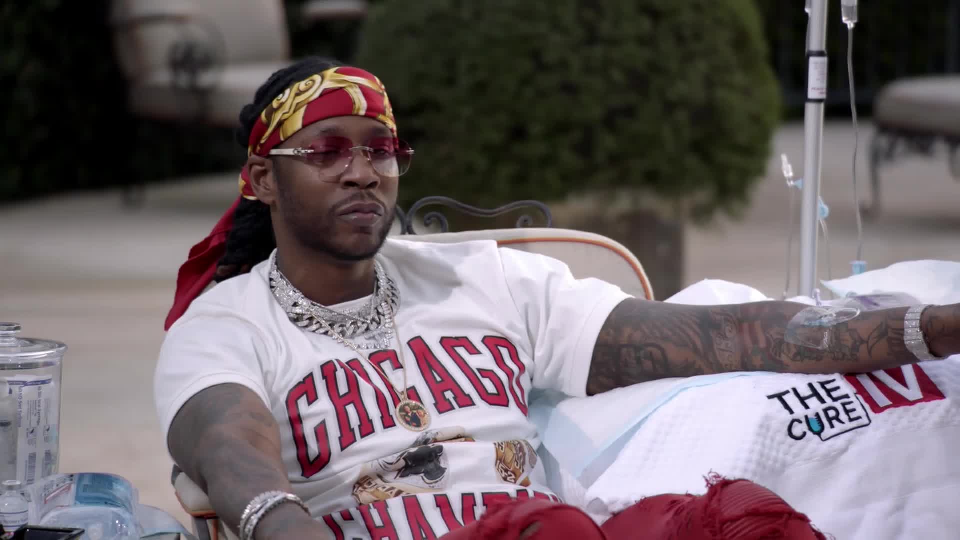 A man in a white "CHICAGO" t-shirt and red bandana rests with an IV drip in his arm. A white blanket with "THE CURE IV" printed on it is spread beneath him.