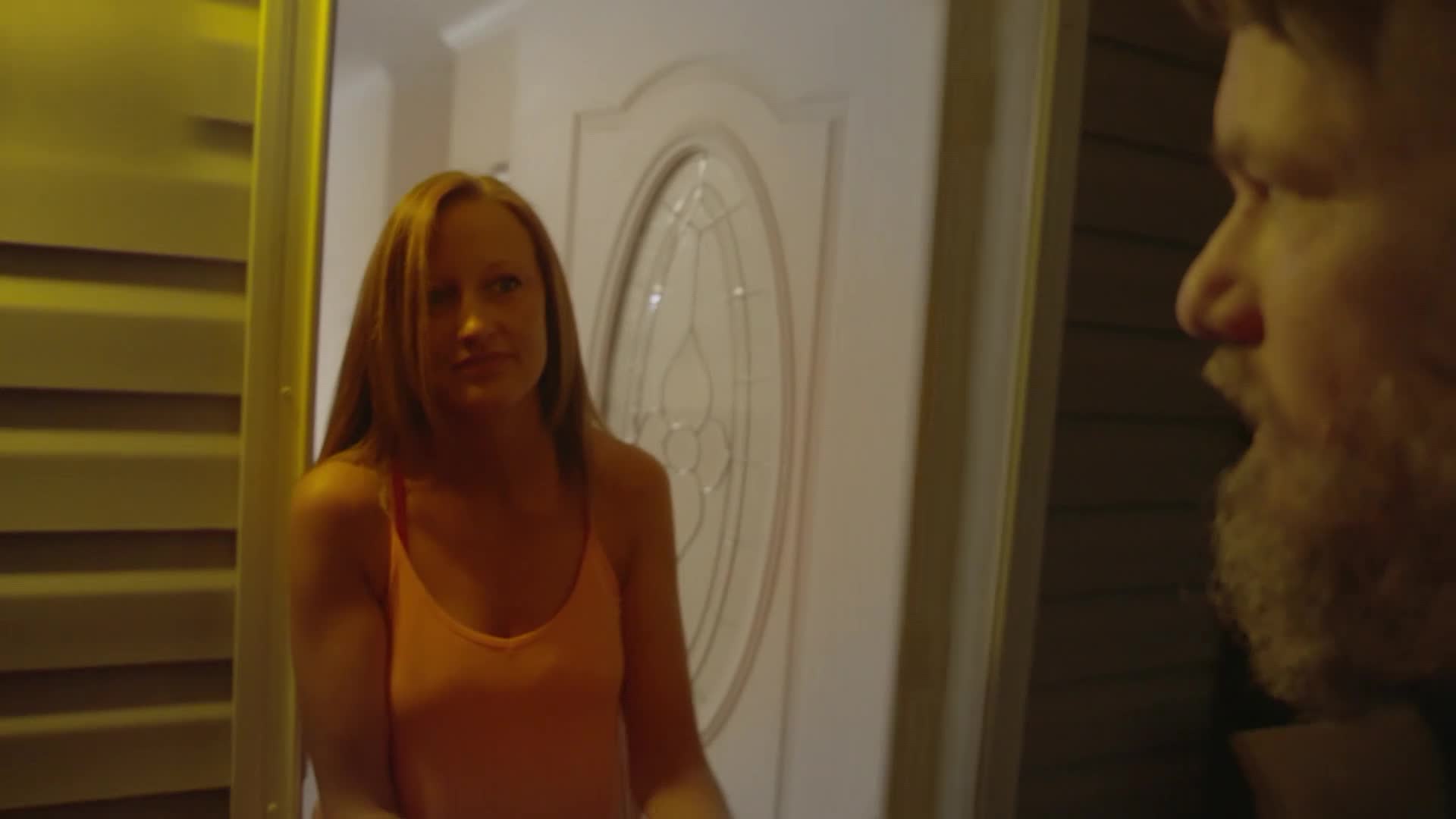 A woman in an orange tank top stands by a door, looking off to the side. A man's bearded face is in the foreground, his mouth slightly open.