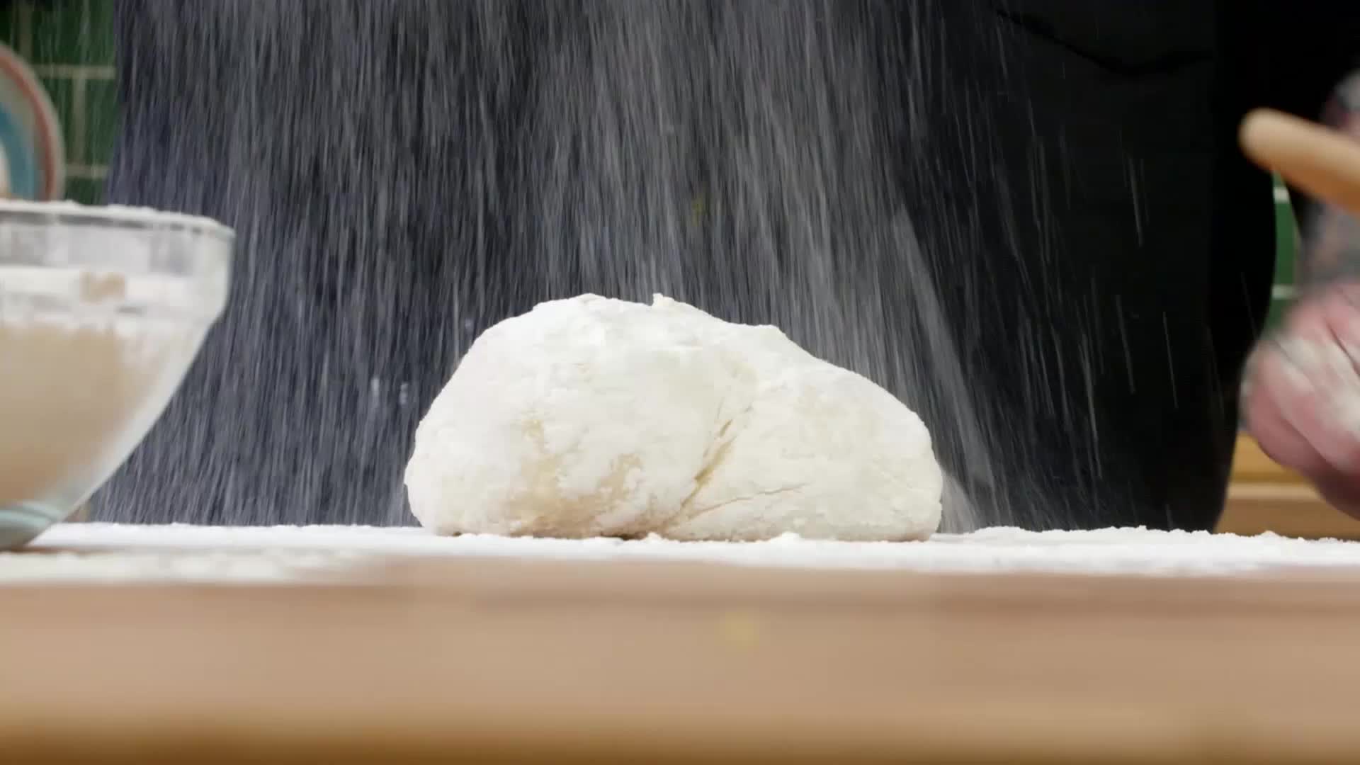 Flour falls like snow onto a mound of dough. A hand, dusted white, grips a wooden rolling pin.