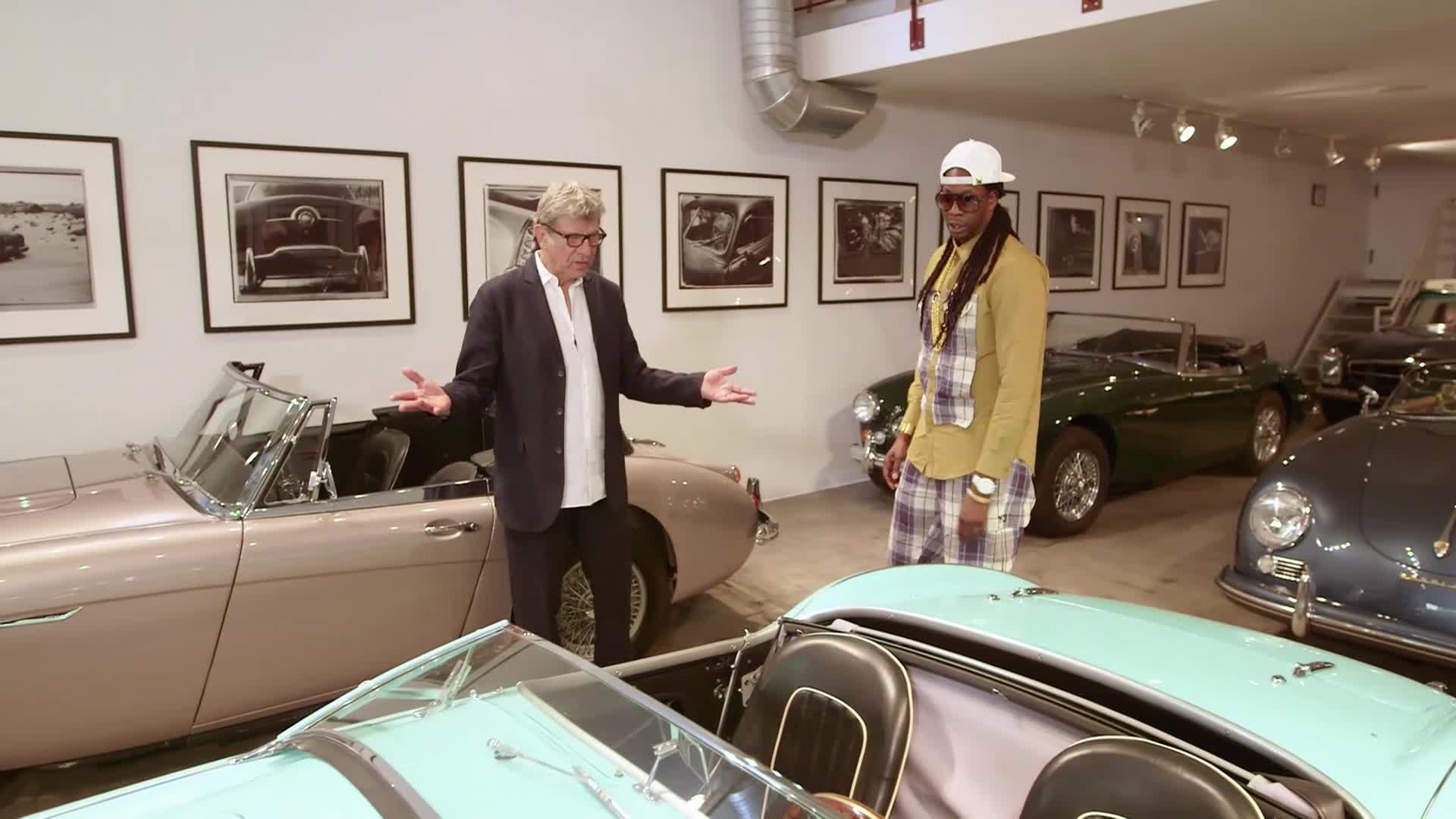 Two men stand in a garage filled with vintage convertibles, one gesturing broadly as if explaining something. The other, dressed in a distinctive yellow shirt and plaid pants, listens intently.