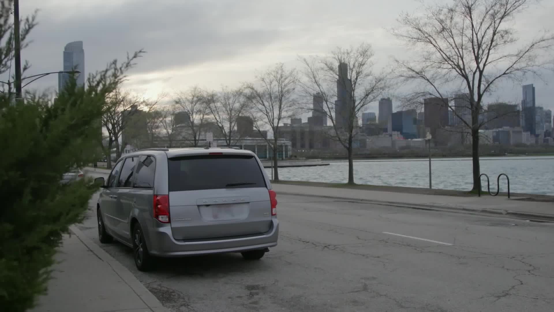 A silver minivan sits parked on the side of a road overlooking Lake Michigan, with the Chicago skyline in the distance. The overcast sky hangs heavy, typical for a day in the United States.