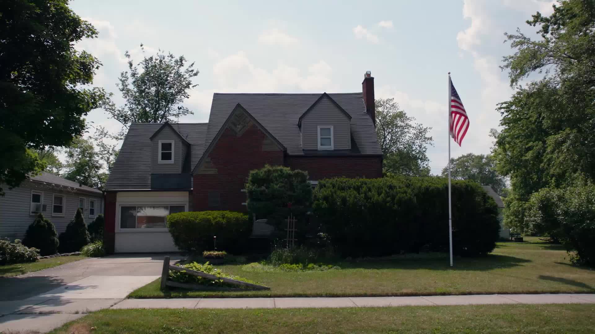 An American flag waves from a tall pole in front of a brick house. The flag's stripes are crisp against the bright sky.