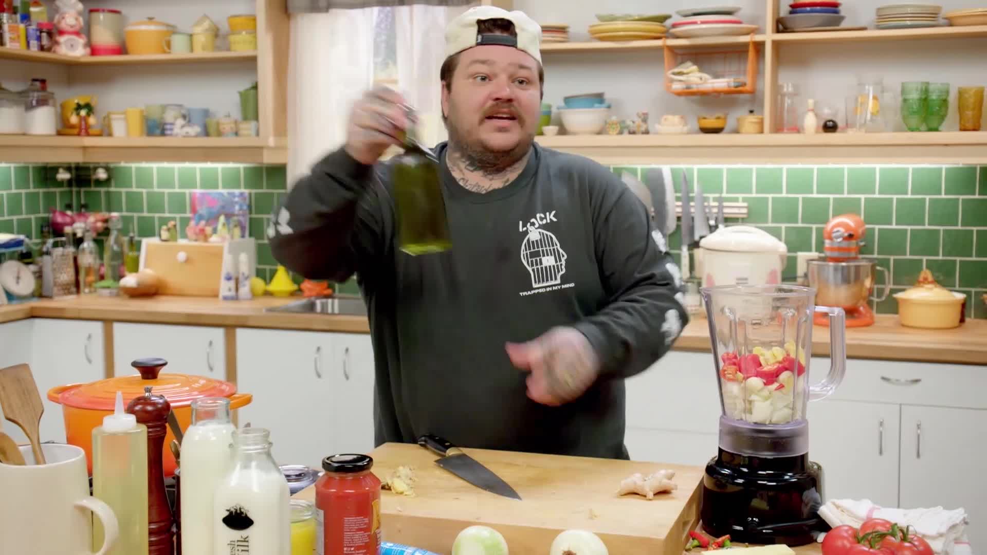 A man in a kitchen holds up a bottle of olive oil, about to pour it into a blender filled with chopped vegetables. He's on the set of a VICE TV cooking show.