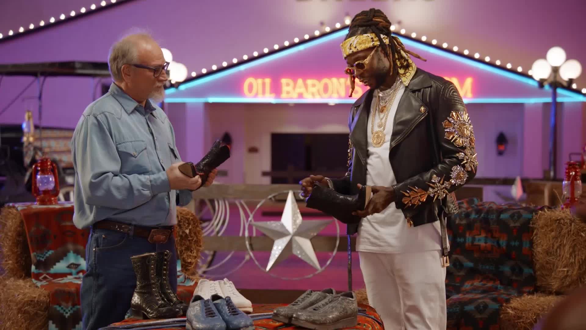 An older man in a denim shirt holds up a dark leather boot. Across from him, a man in a flashy jacket examines a similar boot.