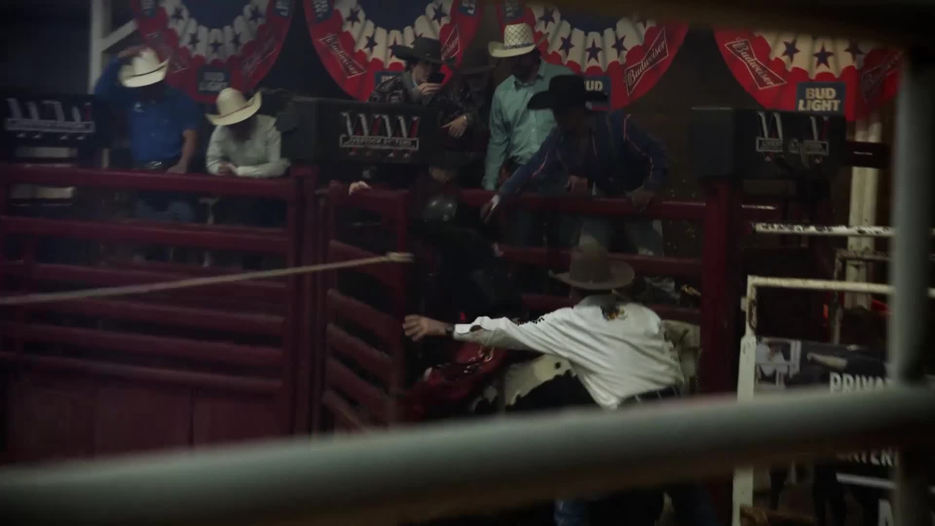A cowboy in a white shirt is reaching into the chute to help another rider. The bull is bucking inside the pen.
