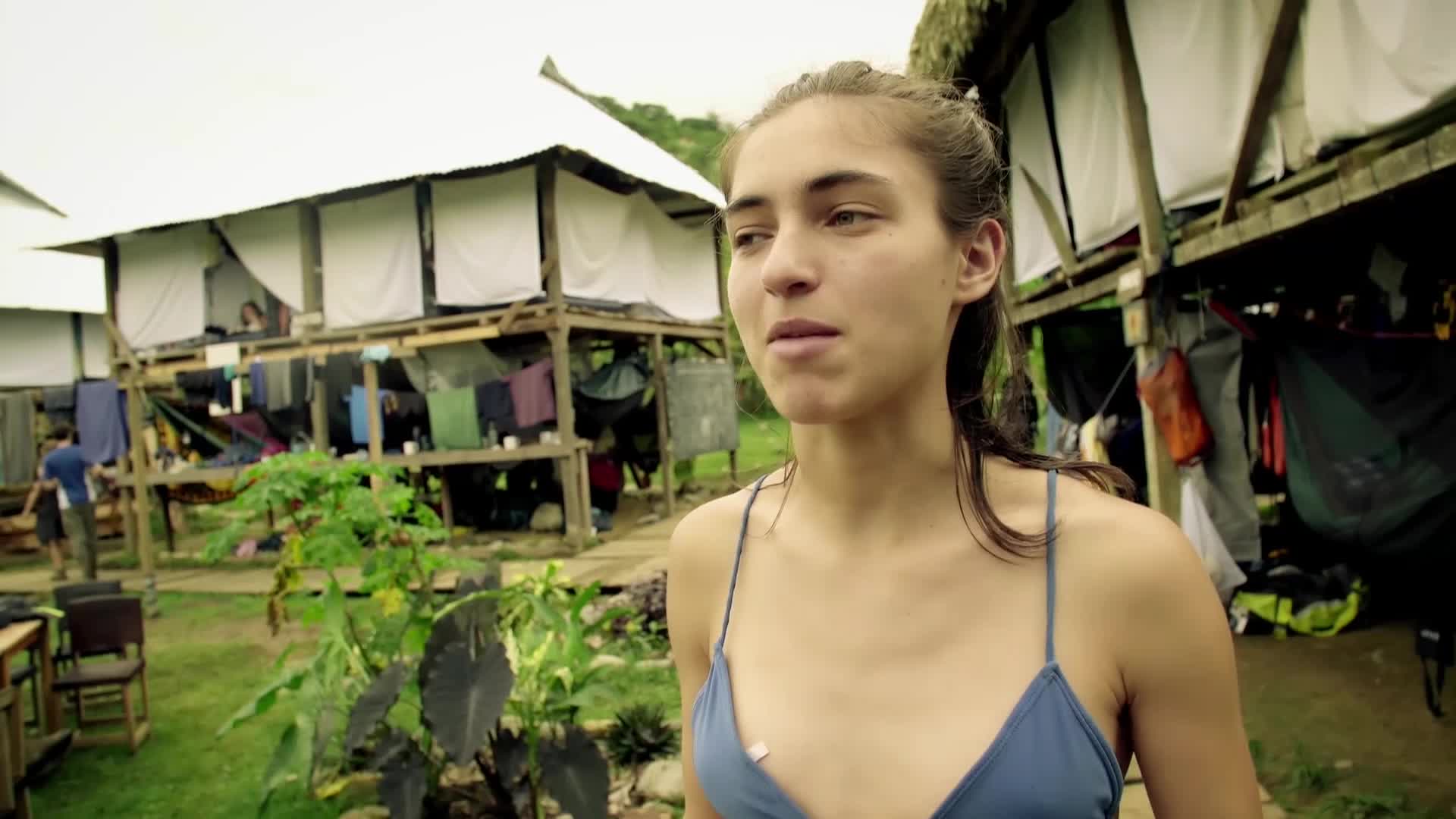 A young woman stands in the foreground, her gaze directed slightly off-camera. Behind her, a rustic compound of wooden structures with thatched roofs houses laundry hanging to dry and people moving about.
