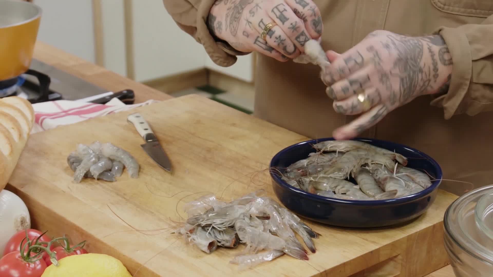 Hands, covered in tattoos, work to peel shrimp over a wooden cutting board. A blue bowl overflows with more of the seafood, ready for preparation.