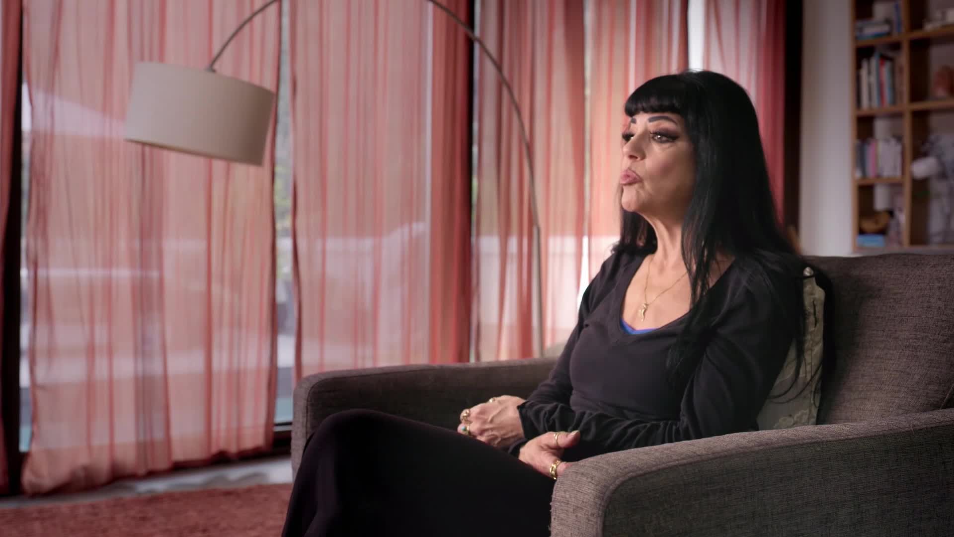 A woman with dark hair and heavy eyeliner sits in a plush armchair, her hands clasped in her lap. Behind her, sheer, coral-colored curtains diffuse the light from a large window.