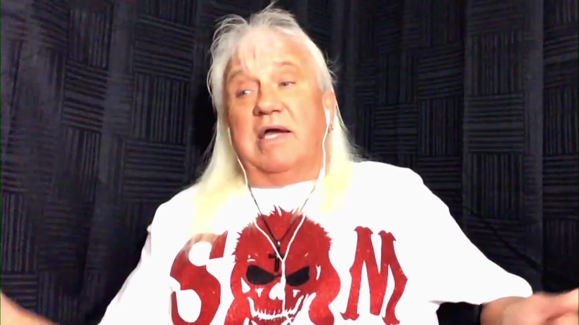A man with long, white hair speaks, his mouth open mid-sentence. He wears a white t-shirt with a large red skull graphic and the word "STORM" across it, the kind of thing you might see on VICE TV.