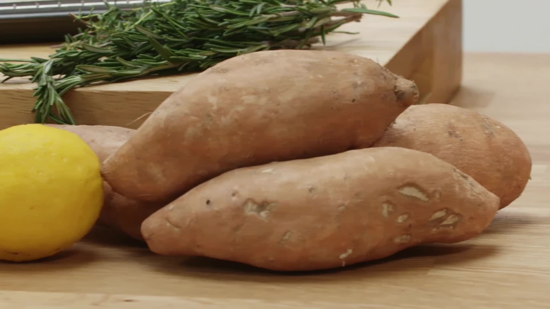 A pile of sweet potatoes sits on a wooden cutting board next to a bright yellow lemon. A sprig of fresh rosemary rests above them, ready for the kitchen.