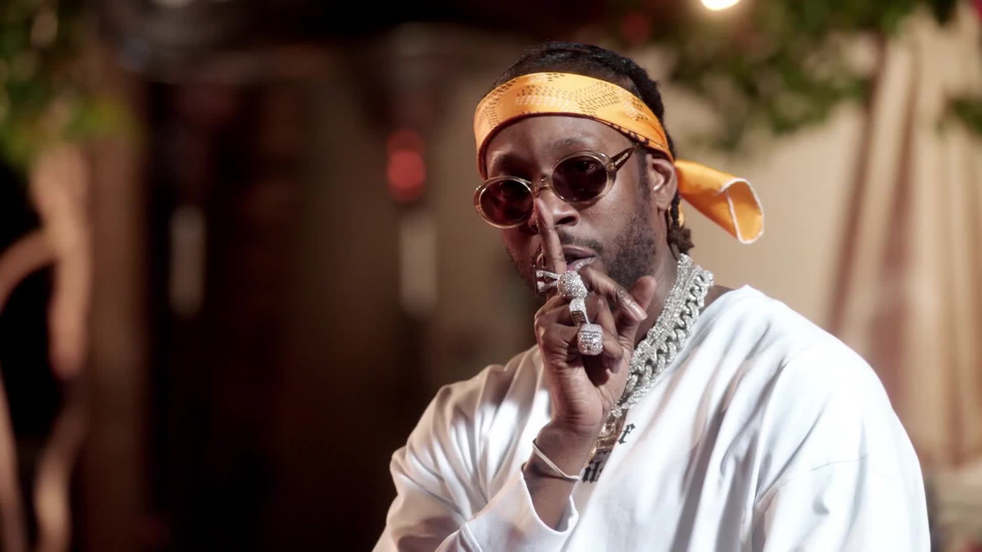 The man in the orange bandana and sunglasses holds a finger to his lips, a gesture of silence. He's wearing a thick, iced-out chain that catches the light.