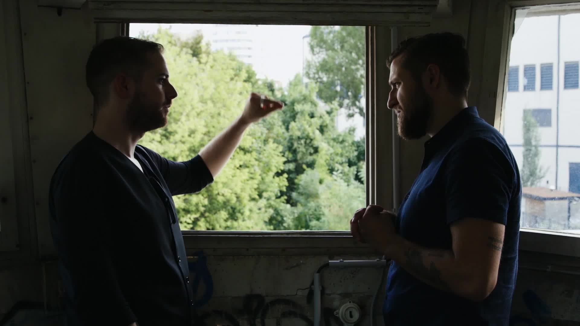 Two men stand in front of a large window, framed by the gritty interior of what looks like an abandoned train car. One man gestures with his hand, explaining something to the other, who listens intently. Outside, lush green trees and a distant building suggest an urban fringe setting.