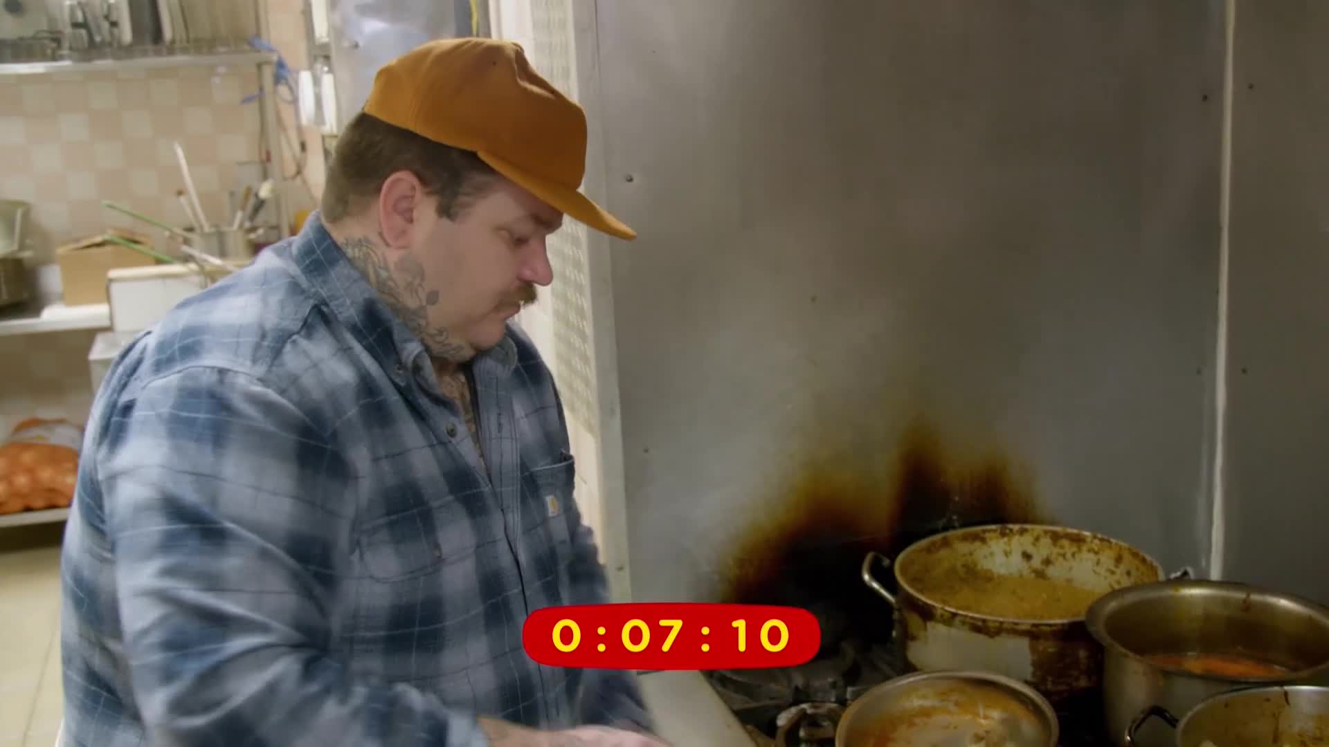 A man in a plaid shirt and a tan hat leans over a stove, his tattoos visible on his neck. Several pots of food sit on the burners, with a timer showing 0:07:10.