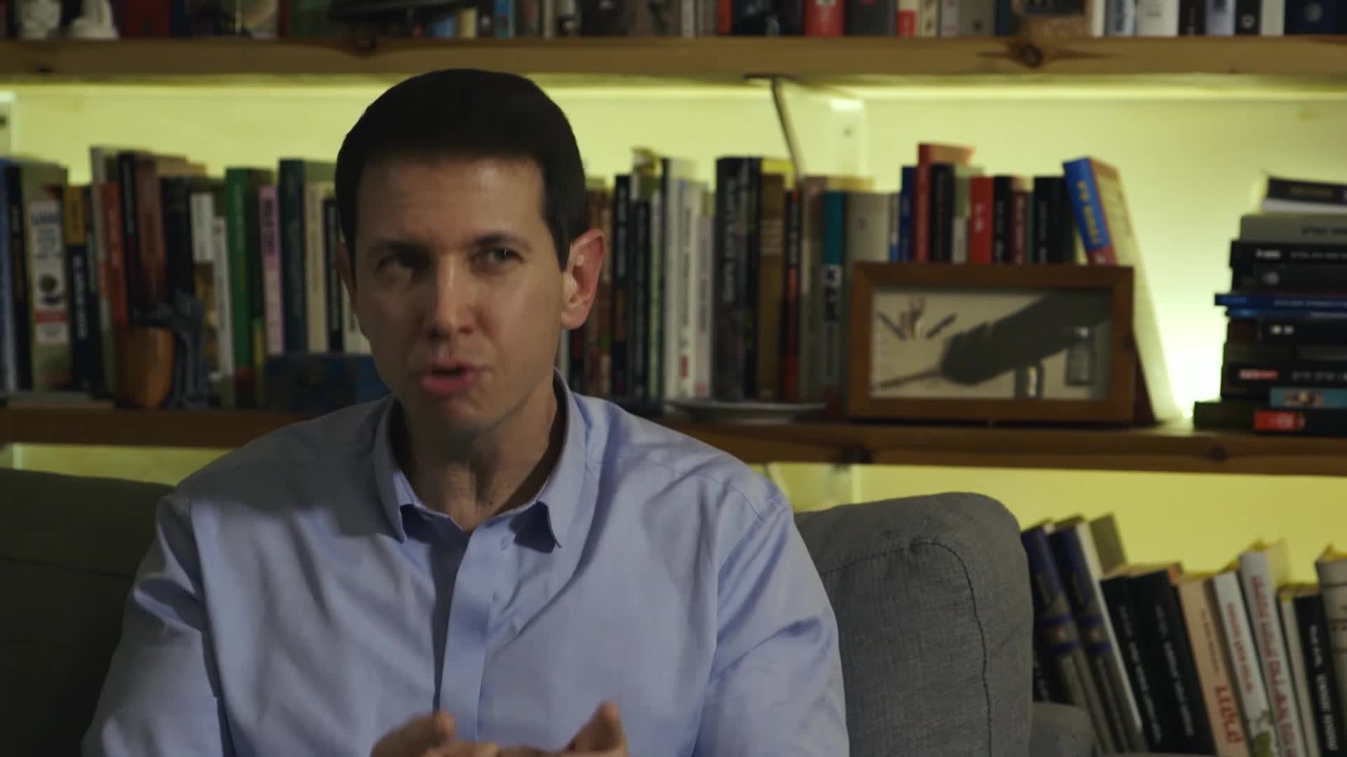 A man in a blue button-down shirt is speaking, his hands gesturing slightly. Behind him, shelves packed with books are illuminated by a warm light.