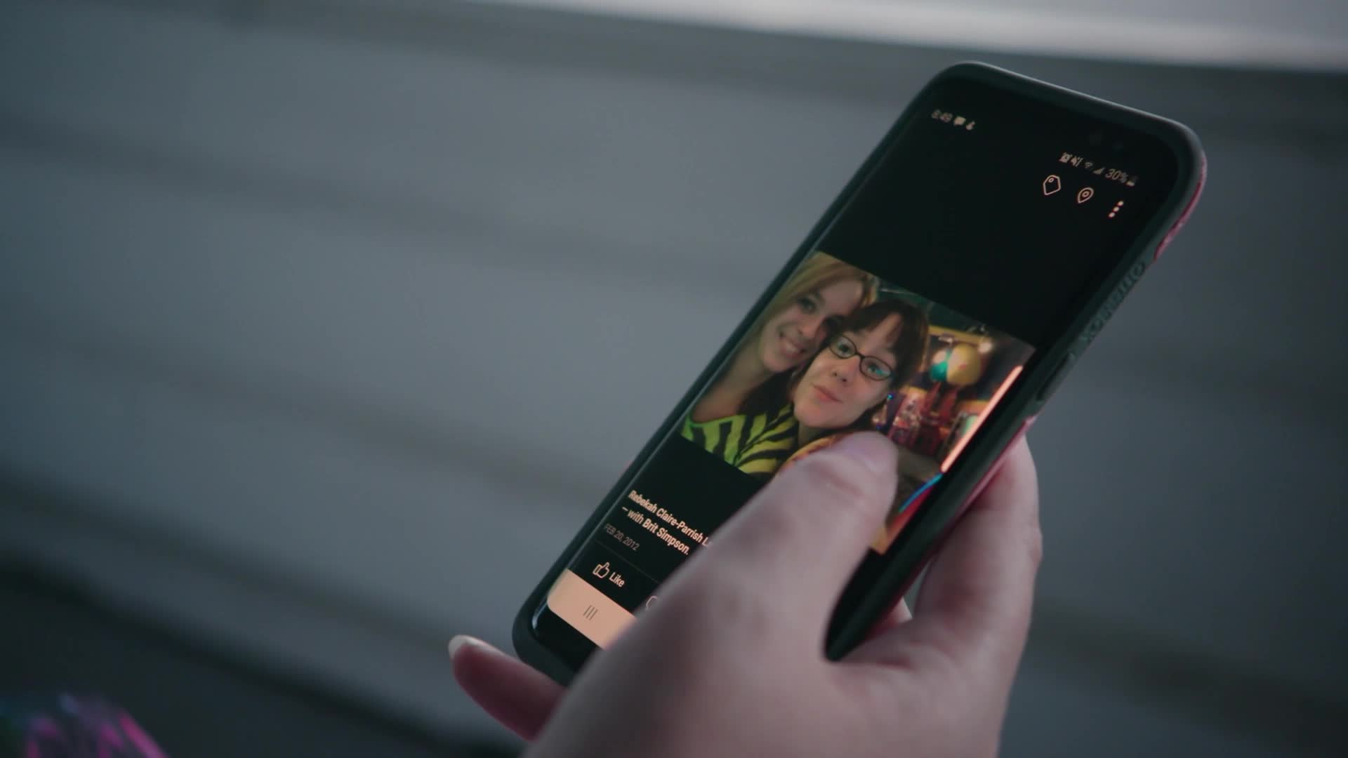 A thumb scrolls through a Facebook post on a smartphone, revealing a selfie of two smiling women. The screen displays their names and a date, hinting at a personal memory shared online.