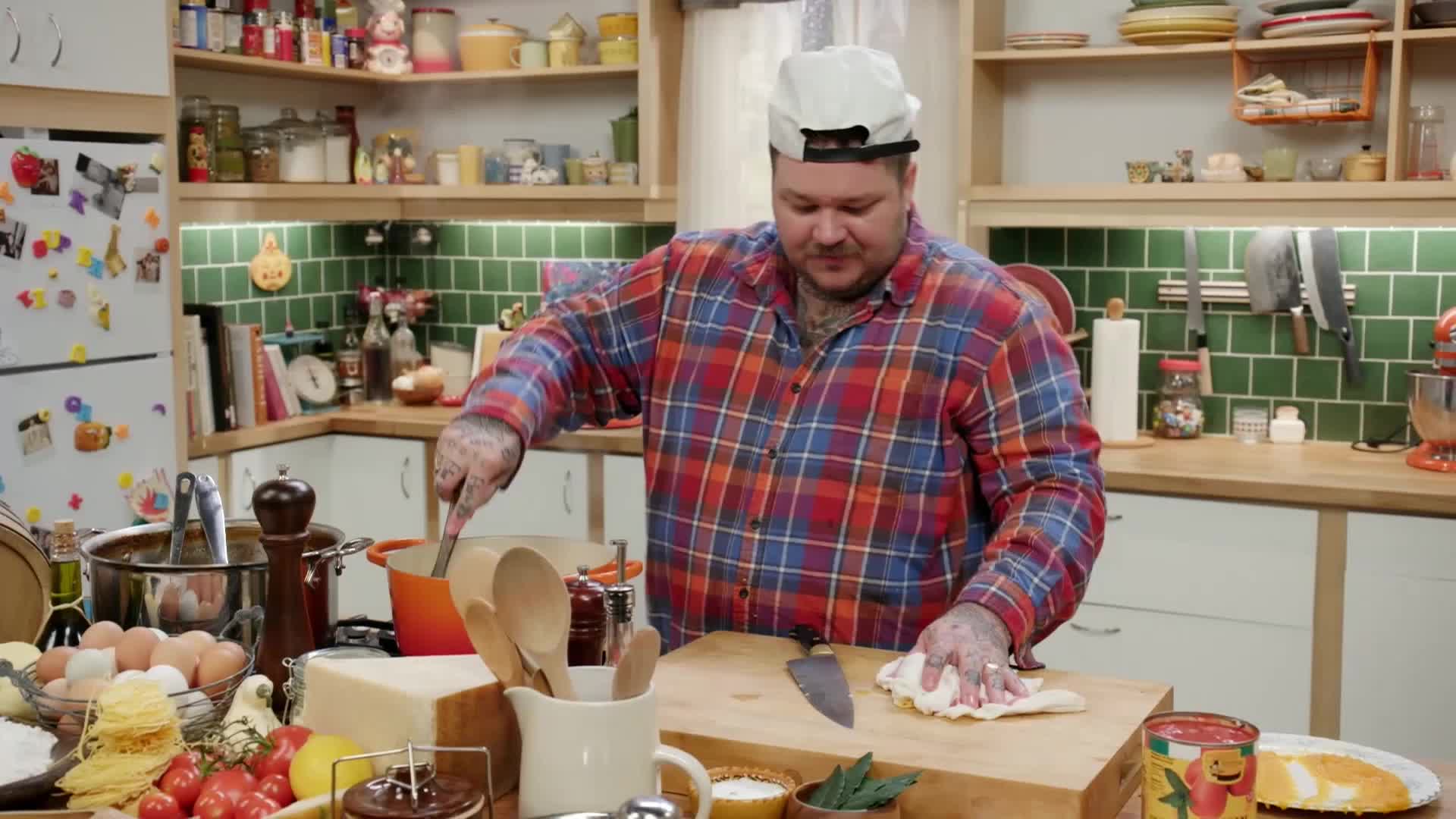 A man in a plaid shirt stirs a pot on the stove, steam rising. He then turns to pat down a piece of dough on a cutting board.
