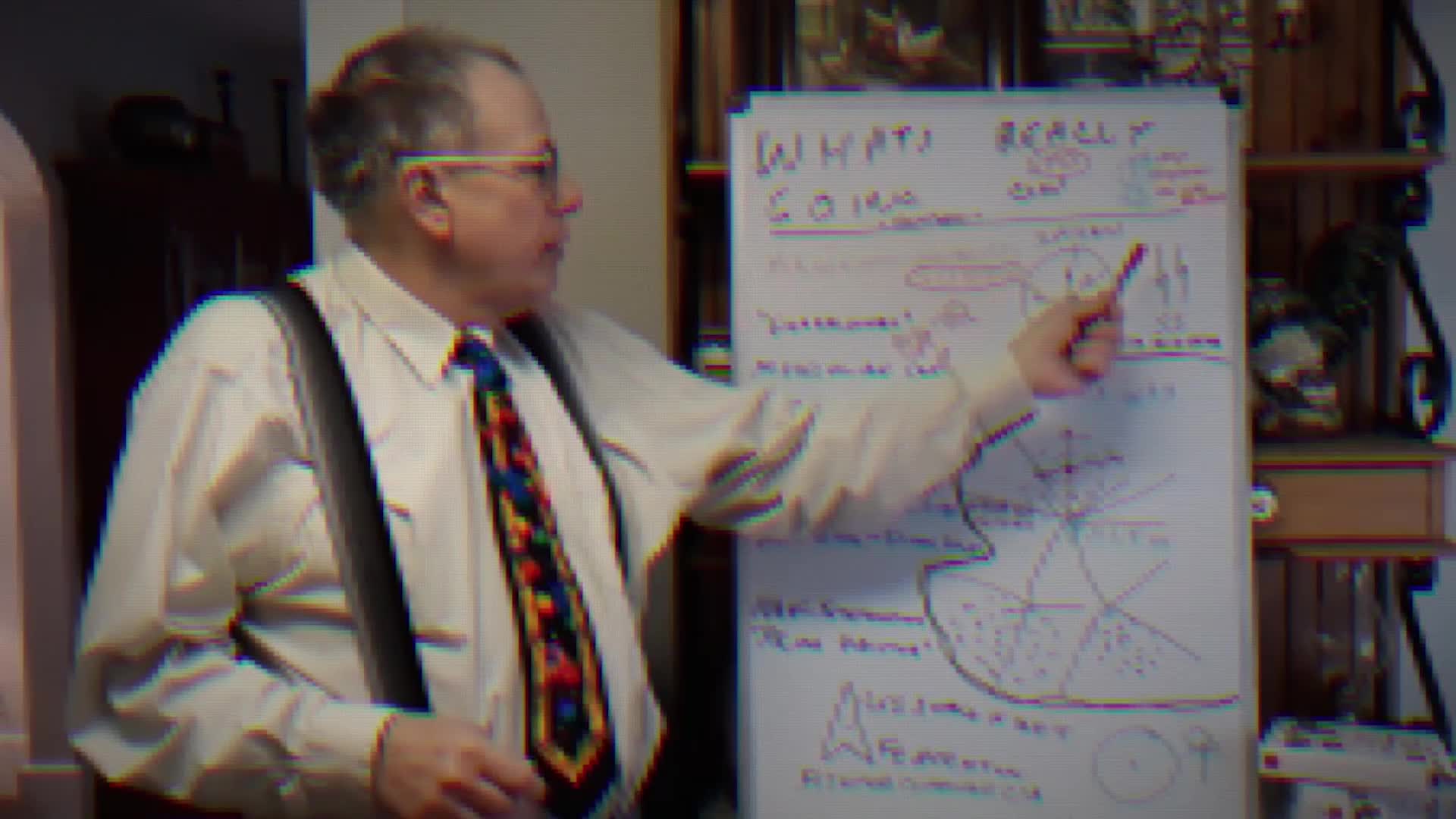 A man in a collared shirt and suspenders points a red pen at a whiteboard covered in diagrams and text. He's explaining something, his arm extended towards the board.