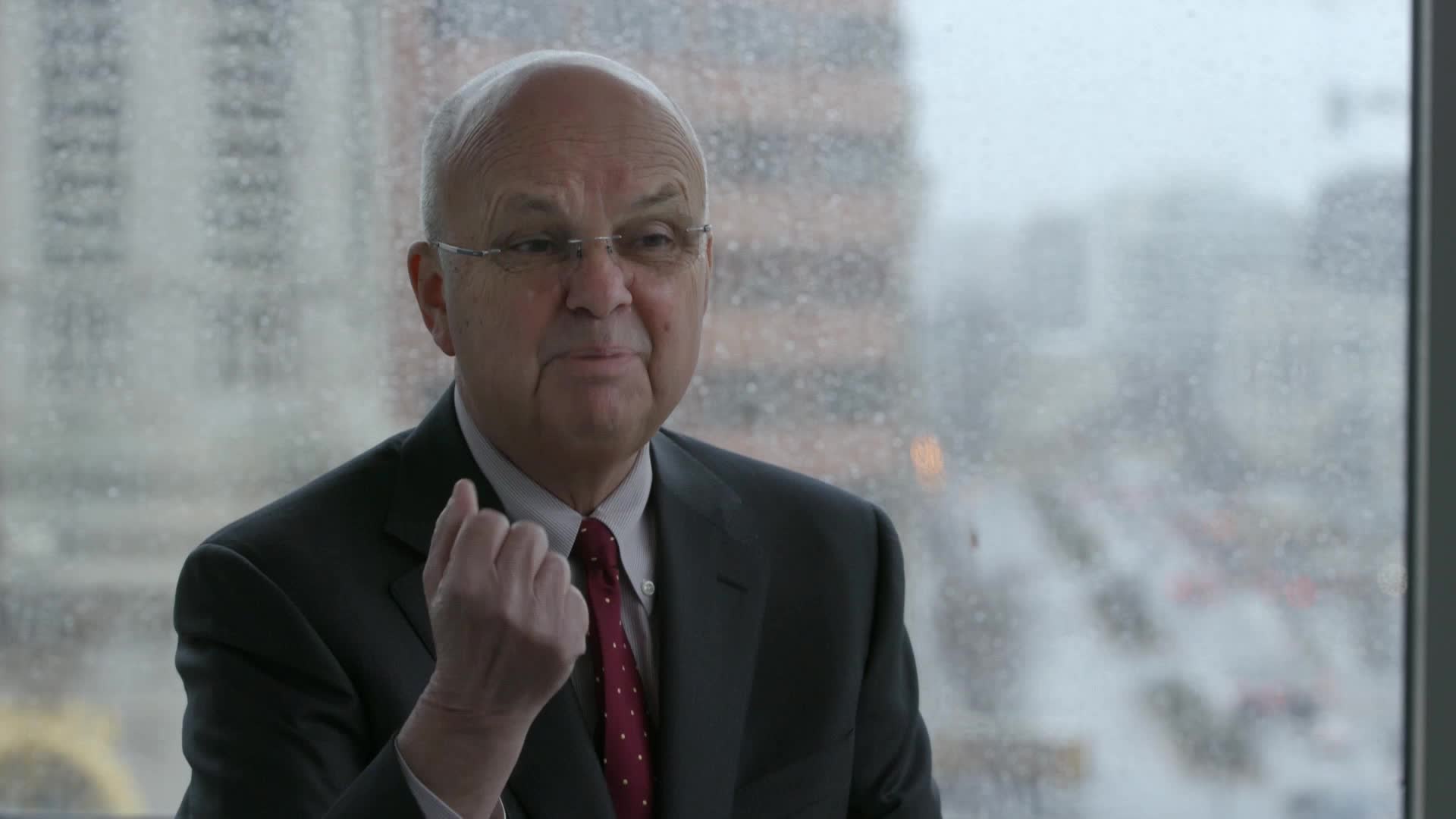 Rain streaks down the window behind a man in a suit, his hand raised as if making a point. The city outside is a muted backdrop, blurred by the weather and distance.