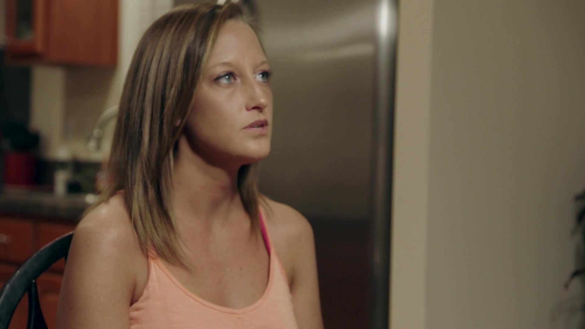 A young woman with blonde hair, wearing an orange tank top, sits in a kitchen. Her eyes are fixed on something to her right, her expression thoughtful.