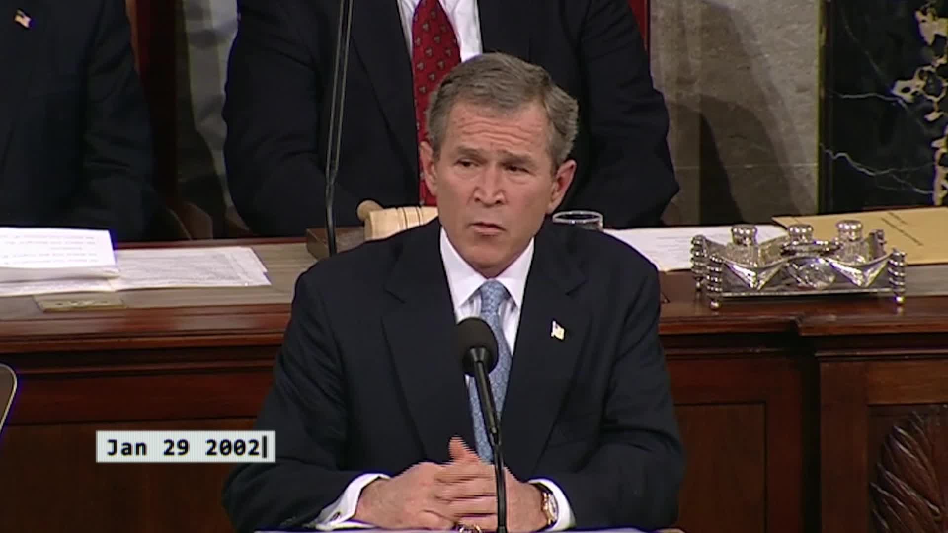 The President speaks at the podium, his hands clasped before him. Papers sit on the polished wood desk.