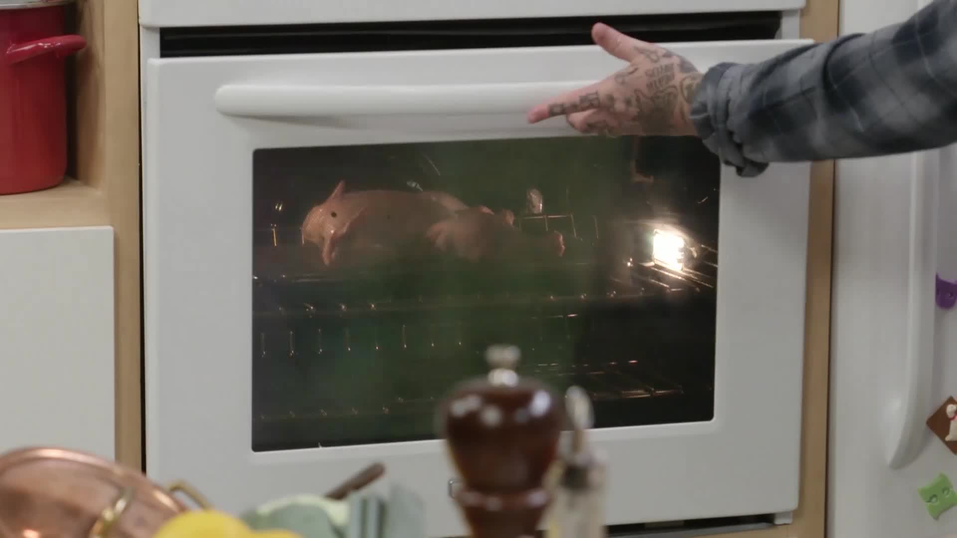 A tattooed hand opens the oven door, revealing a whole pig roasting inside. The bright interior light illuminates the cooking meat.