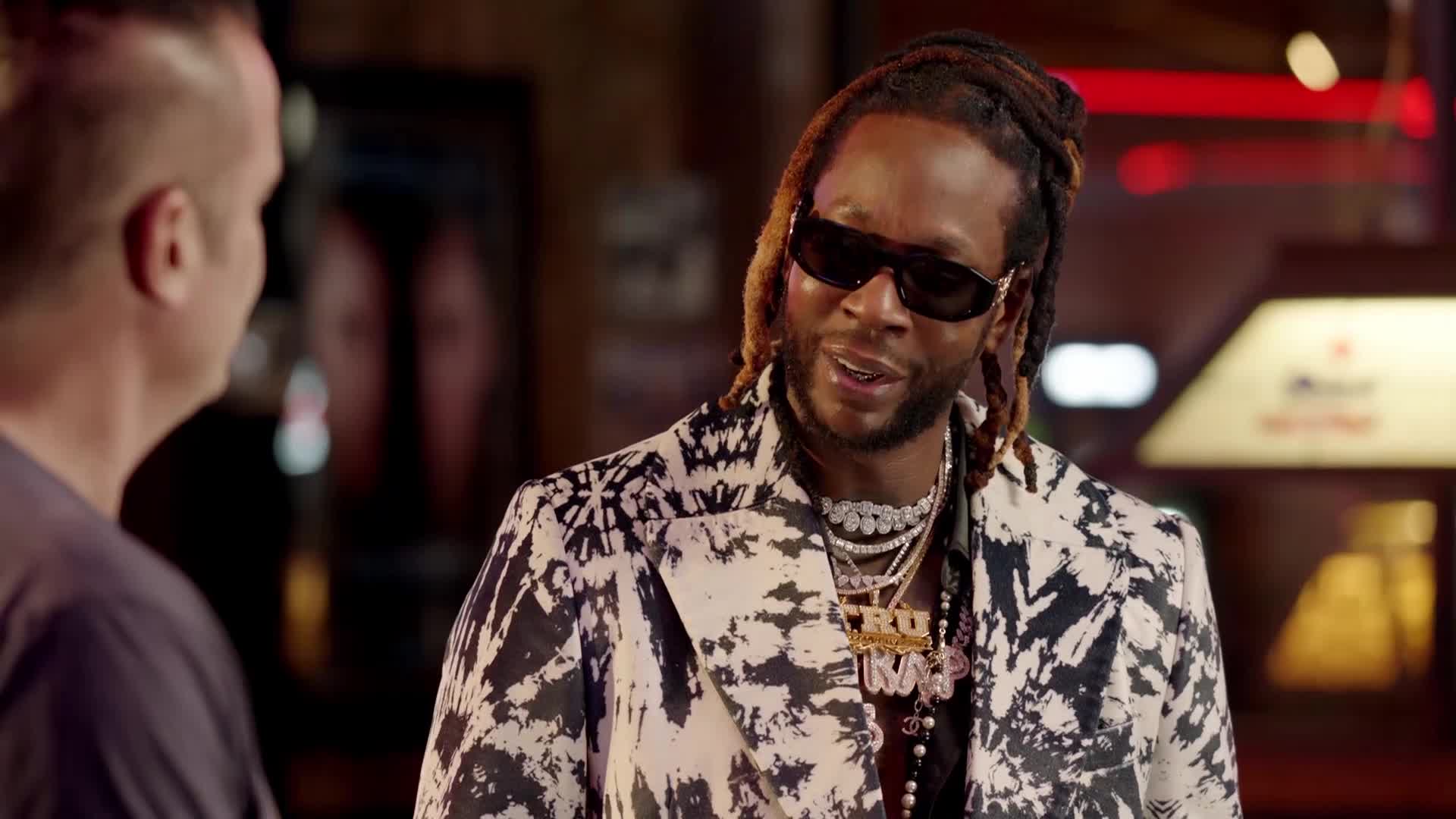 A man with dreadlocks and sunglasses, wearing a patterned jacket and heavy chains, speaks to someone off-camera. He's in a dimly lit room, possibly a bar, with a Heineken sign visible in the background.