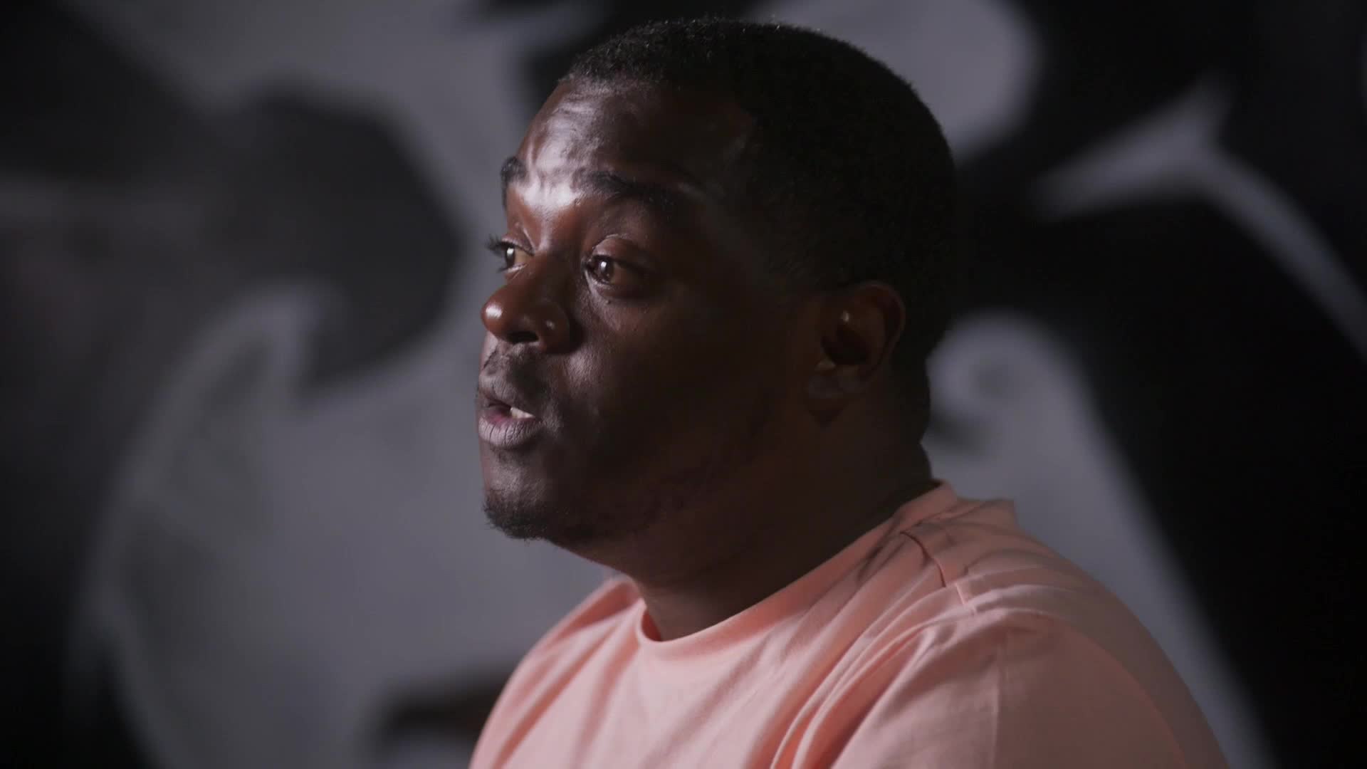 A man in a peach t-shirt speaks, his face illuminated by a light source off-camera. Behind him, a large, abstract mural in black and white looms.