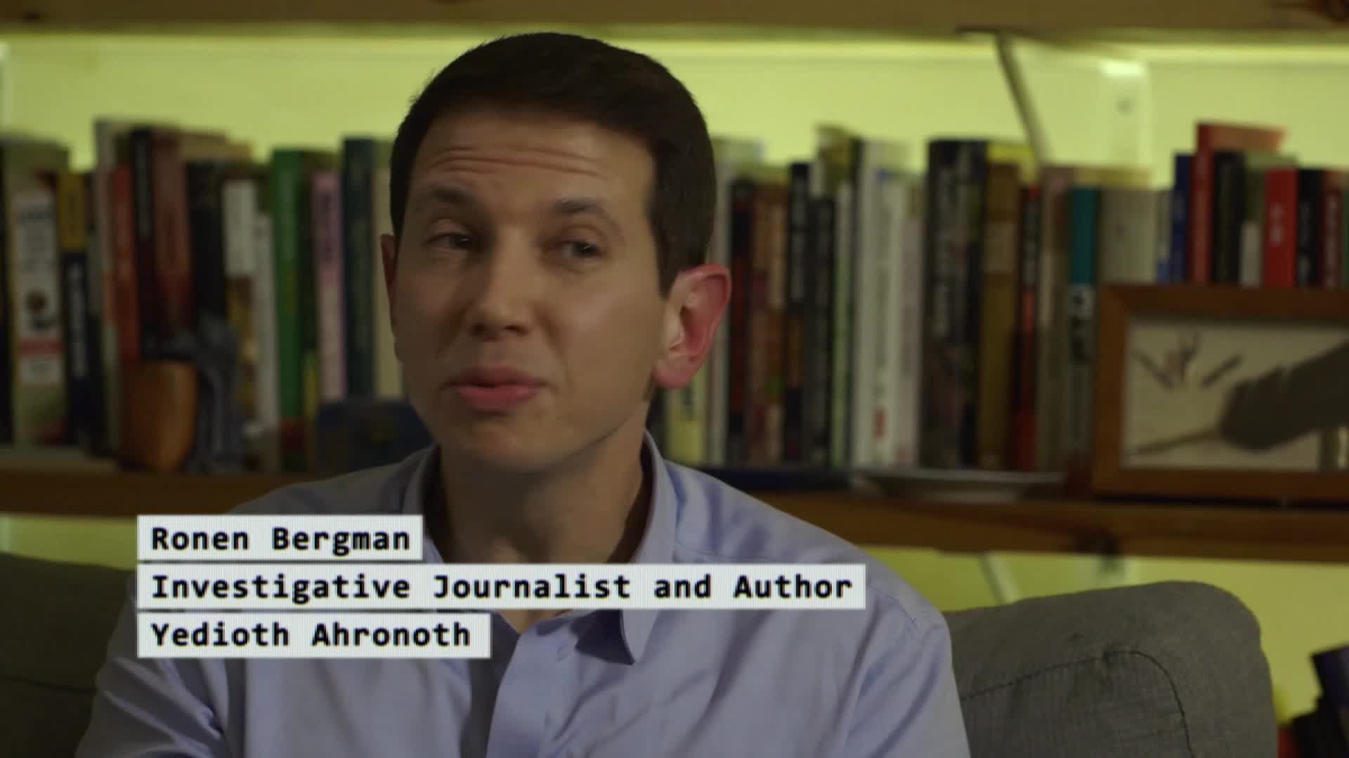 A man with dark hair, Ronen Bergman, speaks directly to the camera. Behind him, shelves packed with books and a framed drawing create a backdrop.