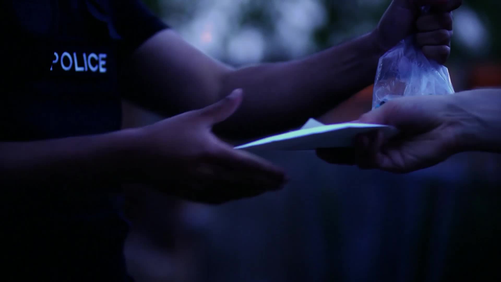 A police officer hands an envelope to another person. The officer's dark shirt has "POLICE" in white letters.