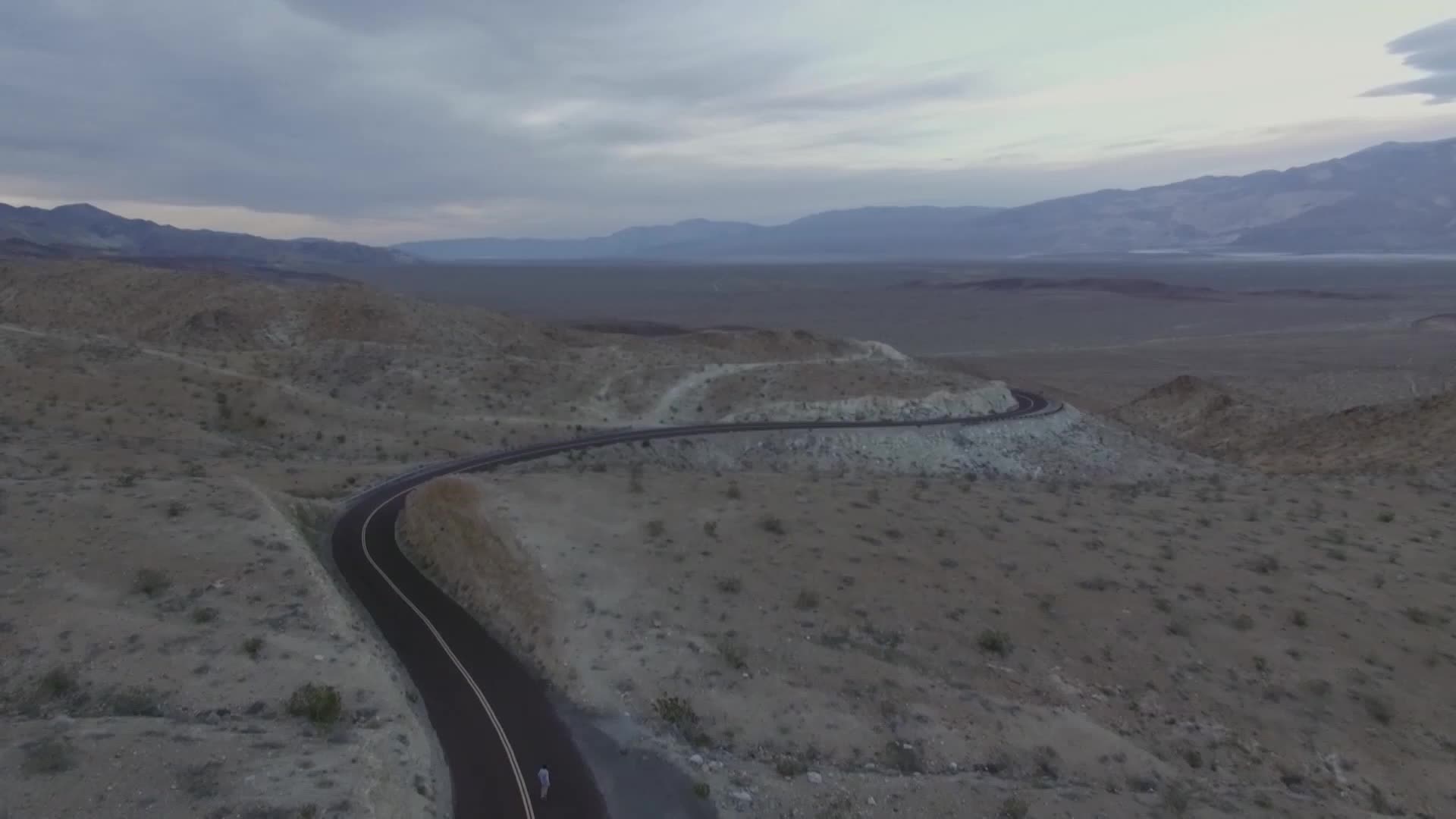 A lone figure on a bicycle pedals along a winding desert highway. The vast, empty landscape stretches out under a cloudy sky, hinting at the isolation of this American journey.