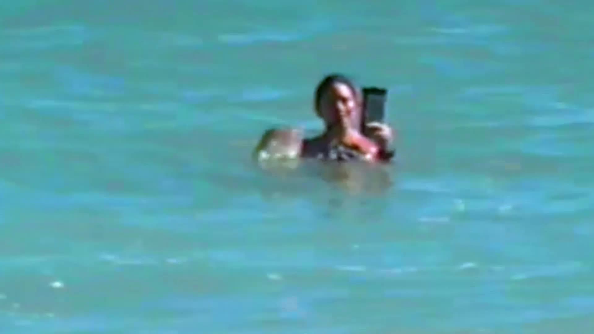 A woman is in the water, holding up a phone to take a selfie. The turquoise water laps around her shoulders.