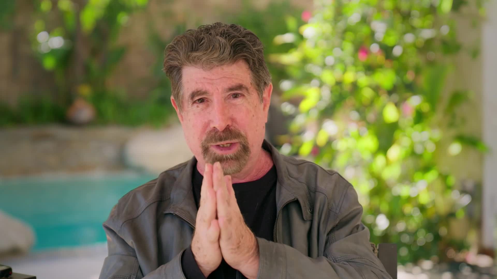 A man with a goatee and salt-and-pepper hair clasps his hands together in front of him. He wears a gray jacket over a black shirt, his expression earnest as he speaks. Behind him, a bright blue pool and lush greenery are visible.