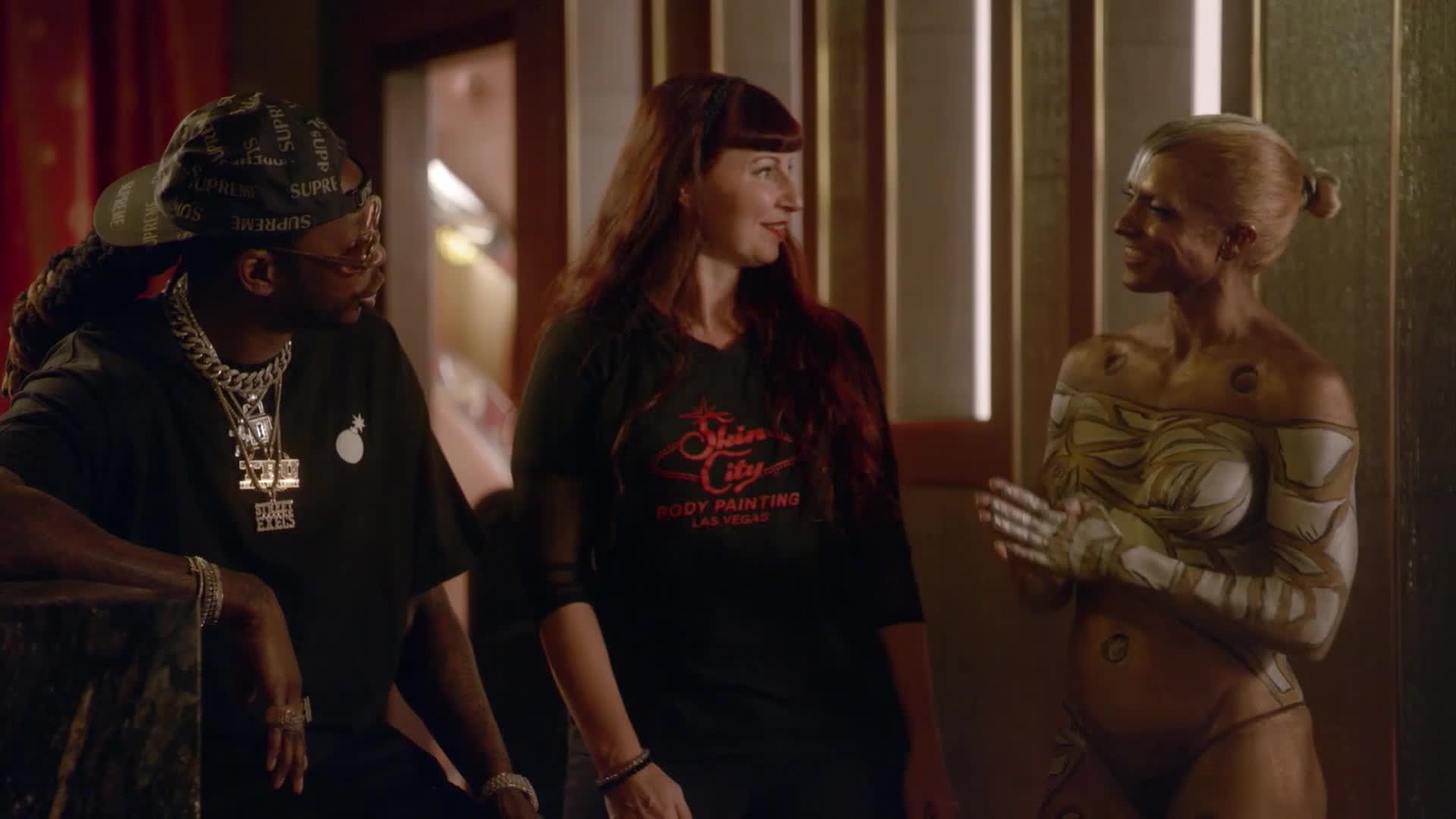 A man in a black t-shirt and a woman in a "Spin City Body Painting Las Vegas" shirt talk with a woman whose body is painted with intricate gold and white patterns. The scene unfolds with the casual atmosphere of a VICE TV production.