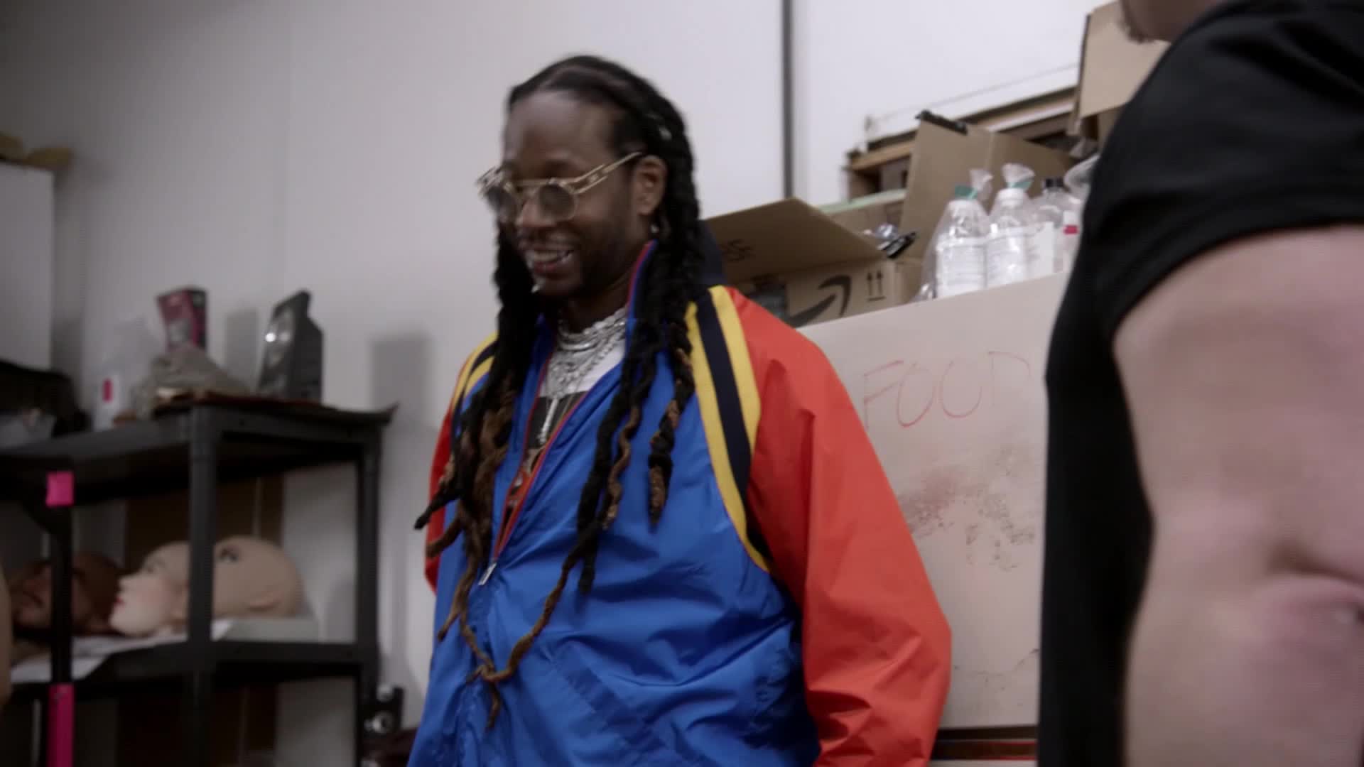 A man with long dreadlocks and glasses smiles, wearing a colorful jacket. He stands near a cardboard box labeled "FOOD" and a shelf with mannequin heads.