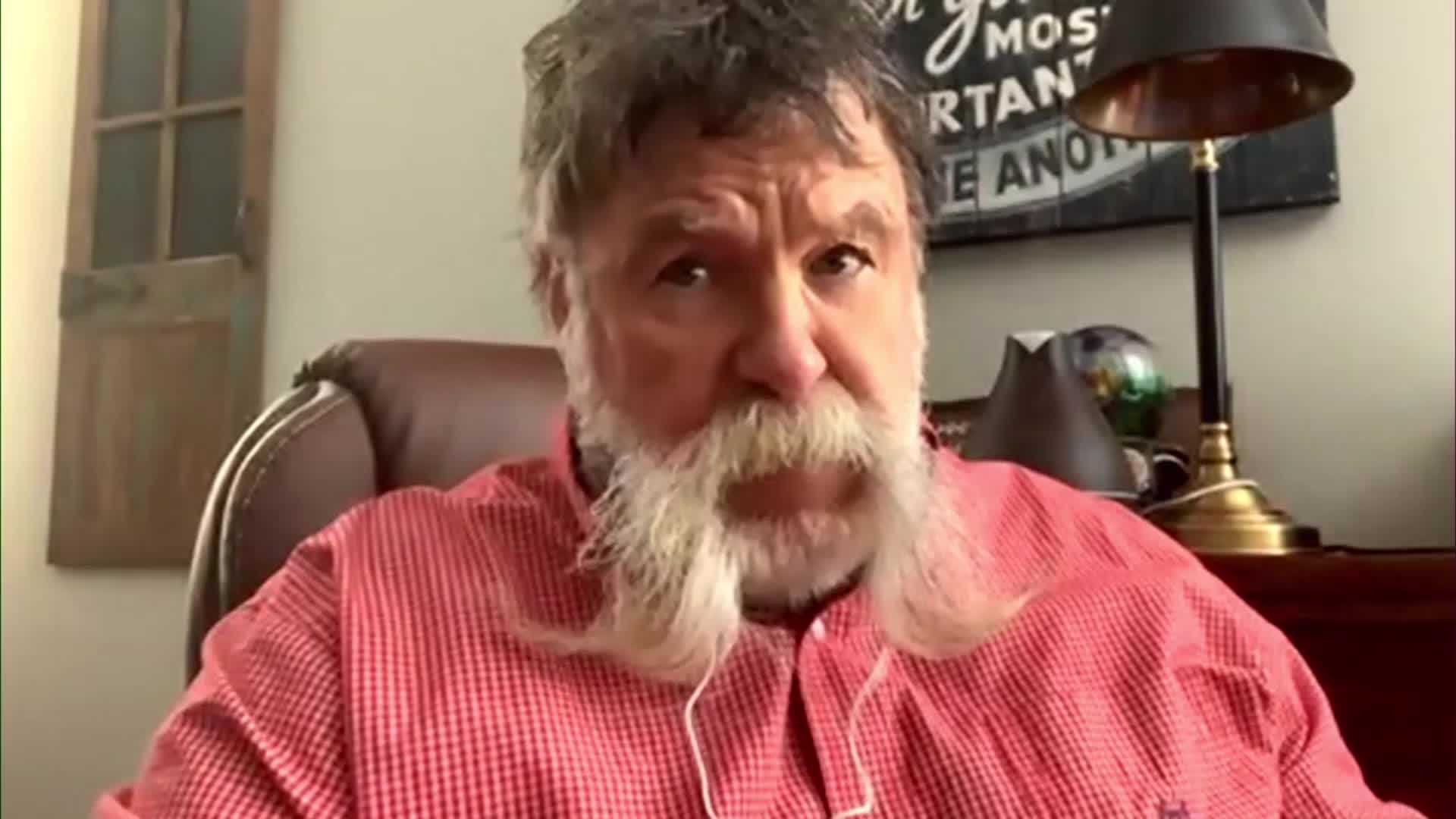 A man with a long, white beard and a red checkered shirt looks directly at the camera. He's seated in a brown leather chair, with a lamp and a framed quote behind him.