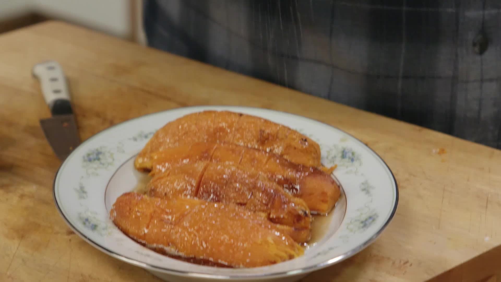 A platter of glazed sweet potato halves sits on a wooden cutting board, a chef's knife resting nearby. This looks like something from a VICE TV cooking segment, a comforting dish prepared with care.