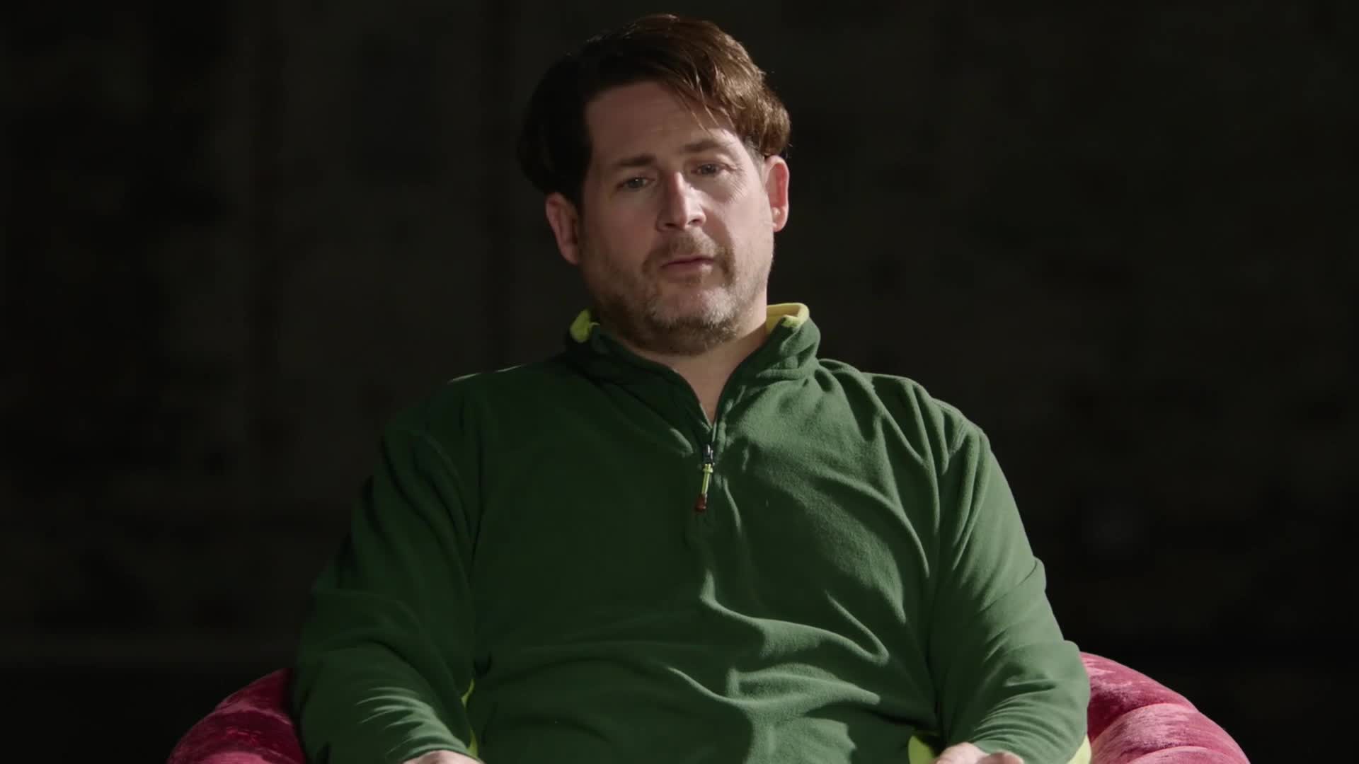A man in a green fleece jacket sits in a chair, looking directly ahead. He has a slight stubble and his hair is styled in a way that suggests a receding hairline.