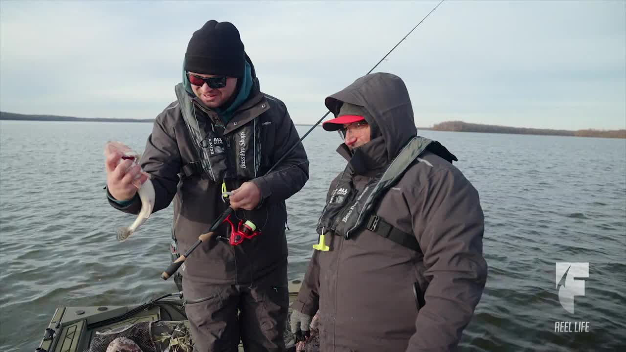 A fisherman in a black beanie holds up a small fish, showing it to his companion on the boat. The other man, bundled in a hooded jacket, looks on with interest.
