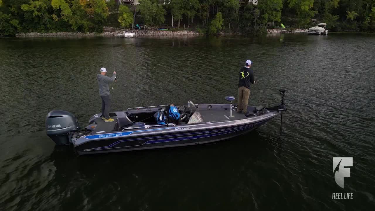 Two anglers, each with a fishing rod, stand on a bass boat, casting lines into the water. The boat, with a Yamaha engine, is moving across the lake, likely in the United States, as shown on the World Fishing Network.

