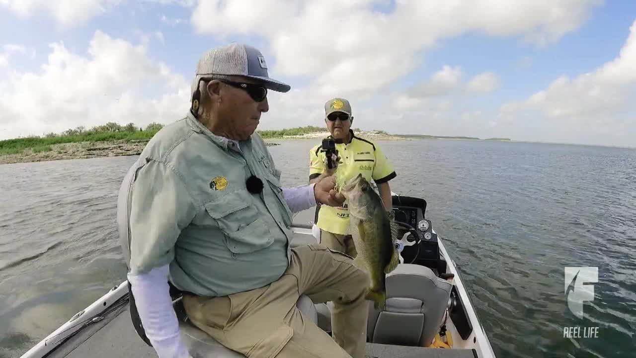 A man in a Bass Pro Shops hat holds up a largemouth bass on a boat, while another man films the catch. The scene unfolds on a sunny day with a calm lake and a distant shoreline.
